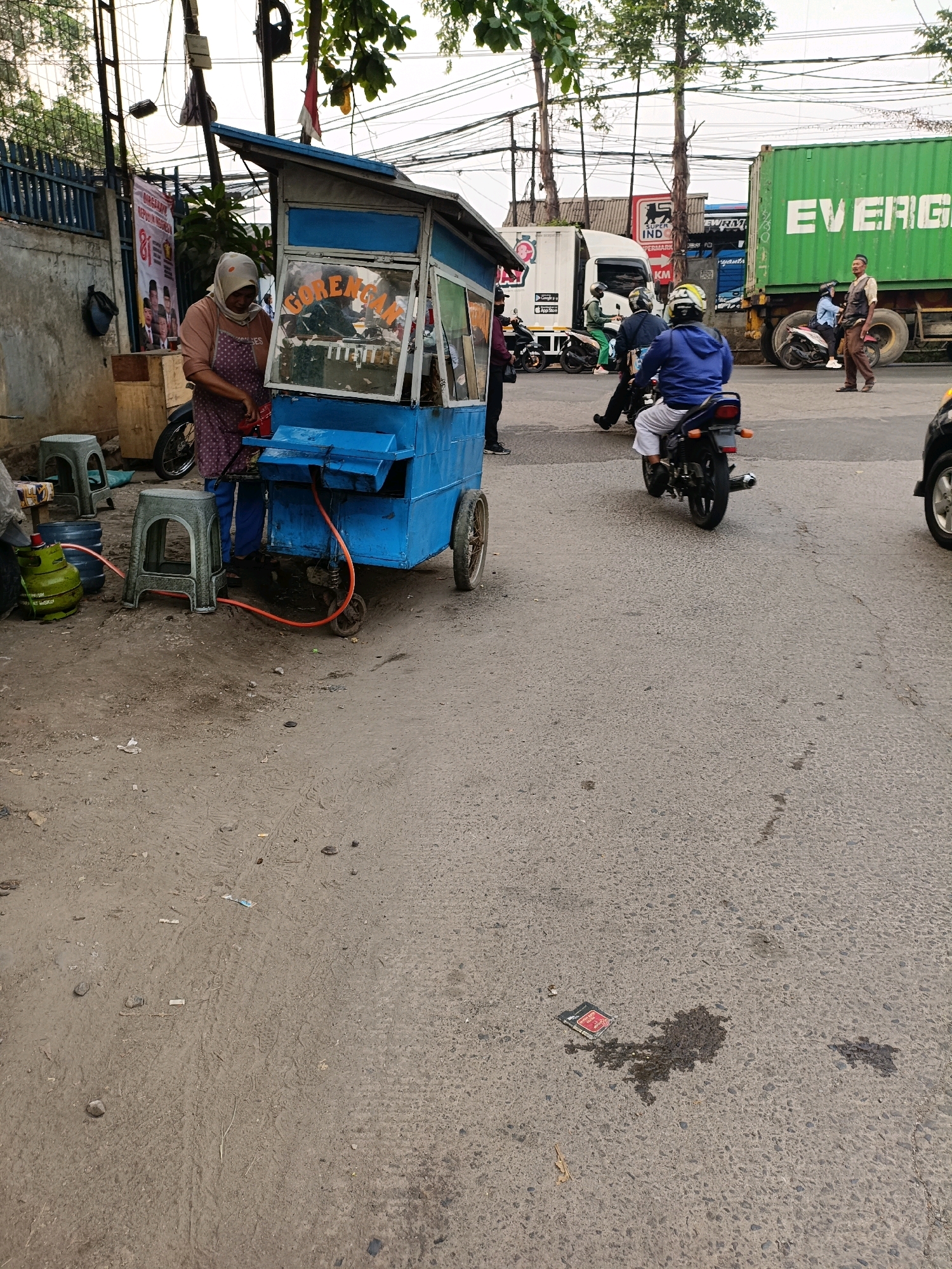 The image captures a street scene in Tangerang, Indonesia, likely near a busy road. In the foreground, a woman wearing a headscarf and apron stands behind a blue food cart, which has "GORENGAN" (Indonesian for fried snacks) written on its glass front. A green gas cylinder is connected to the cart by an orange hose, and two grey plastic stools are placed nearby. The cart is on wheels and appears to be a mobile food stall.
In the background, several people and vehicles are present. A white truck with "EVERGREEN" logo and "EVERGREEN" text is parked on the right side, and other vehicles, including motorcycles and what appears to be a larger truck or bus, are visible on the road. Some individuals are riding motorcycles, while others are standing. One man, dressed in a vest and trousers, stands near the Evergreen truck, and another man in a blue jacket is riding a motorcycle. The ground is paved with asphalt and appears to be somewhat dirty, with some debris scattered around. The overall impression is of a commercial area with ongoing activity.