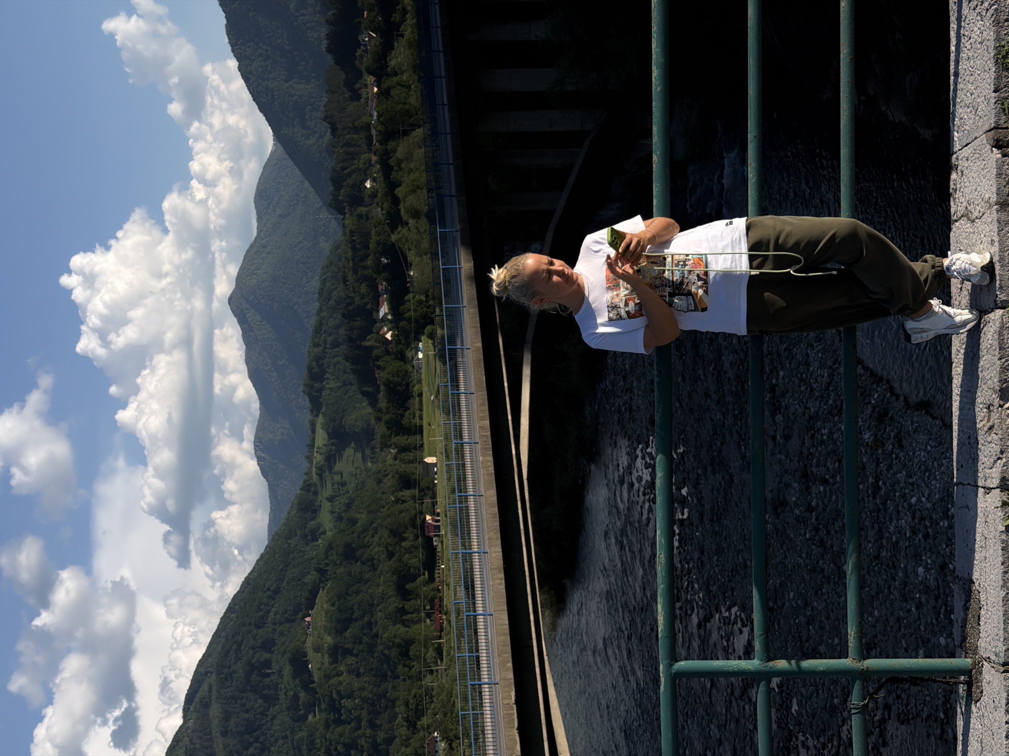 A woman with blonde hair tied up in a bun is standing on the edge of a stone structure, likely a bridge or dam in Mojkovac, Montenegro. She is wearing a white t-shirt with a comic-themed design, olive green sweatpants, and white sneakers. She is holding a mobile phone in her hands and appears to be looking at it. She is leaning against a green metal railing.
Behind her, there is a view of a large dam or bridge structure, with water flowing beneath it. In the background, there are green mountains and a blue sky with cumulus clouds. There are houses and vegetation near the base of the mountains. The overall scene is well-lit with daylight.