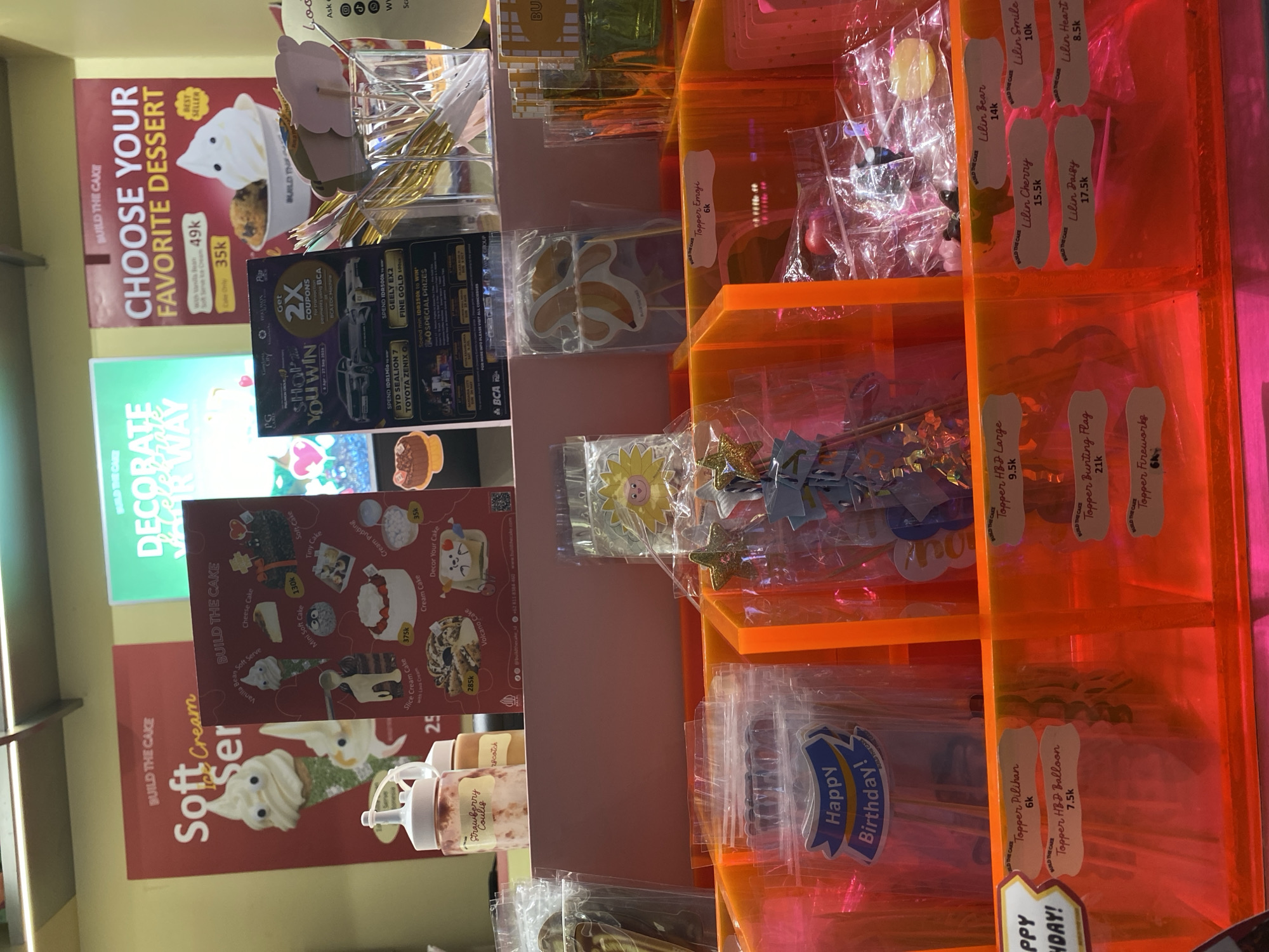 The image depicts a retail display area with various cake decorations and dessert-related items, likely within a bakery or dessert shop. The display is organized into bright orange transparent compartments. In the foreground, there are several plastic-wrapped items, including cake toppers with labels such as "Happy Birthday!", "Topper Pilihan 6K", "Topper HBD Balloon 7.5K", "Topper HBD Large 9.5K", "Topper Bunting Flag 21K", and "Topper Fireworks 6K". There are also other cake decorations and toppings, including small candles labeled "Lilin Bear 14K", "Lilin Smile 10K", "Lilin Cherry 15.5K", "Lilin Daisy 17.5K", and "Lilin Heart 8.5K". In the background, there are promotional posters for a cake shop called "BUILD THE CAKE". One poster advertises "Soft Cream Ser" with images of ice cream cones. Another poster promotes choosing a favorite dessert, showcasing soft-serve ice cream and a cookie. A third poster is a promotional flyer for a "SHOP & WIN" event, featuring cars and prize details, including "GET 2X COUPONS". The overall environment is well-lit and appears to be a clean, commercial setting. The context of the promotional materials suggests the business is located in Indonesia, specifically referencing "BCA" which is a major Indonesian bank, and the prices are in Indonesian Rupiah (indicated by "K" for thousand). The specific location within South Jakarta, Indonesia, cannot be determined solely from the visual information provided.