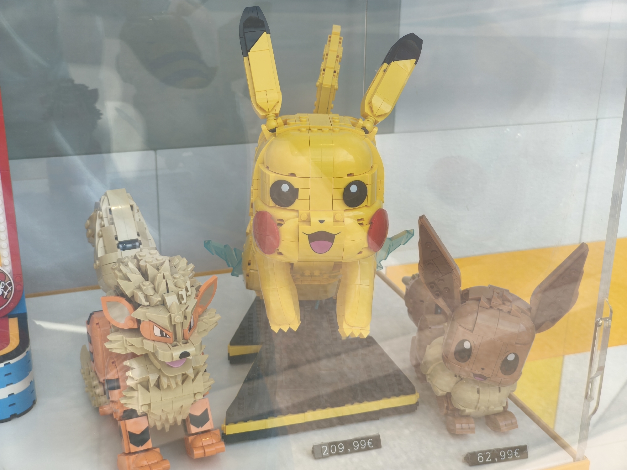 In Egaleo, Greece, this image shows a display window featuring three Pokémon figures constructed from LEGO bricks. From left to right, the figures are Arcanine, Pikachu, and Eevee. Arcanine, the leftmost figure, is primarily tan and orange, with spiky mane and fur details. In the center is a large yellow Pikachu, featuring black-tipped ears, red cheeks, and a wide smile. Behind Pikachu, faint translucent blue LEGO pieces suggest an electrical aura. To the right is a brown Eevee, with large, pointed ears and a fluffy mane. Each figure is displayed on a dark, tiered platform, and price tags are visible below Pikachu and Eevee. A price tag of "209,99€" is under Pikachu, and "62,99€" is under Eevee. The display is behind glass, reflecting the environment and some blurred shapes, possibly of passersby or store interiors.