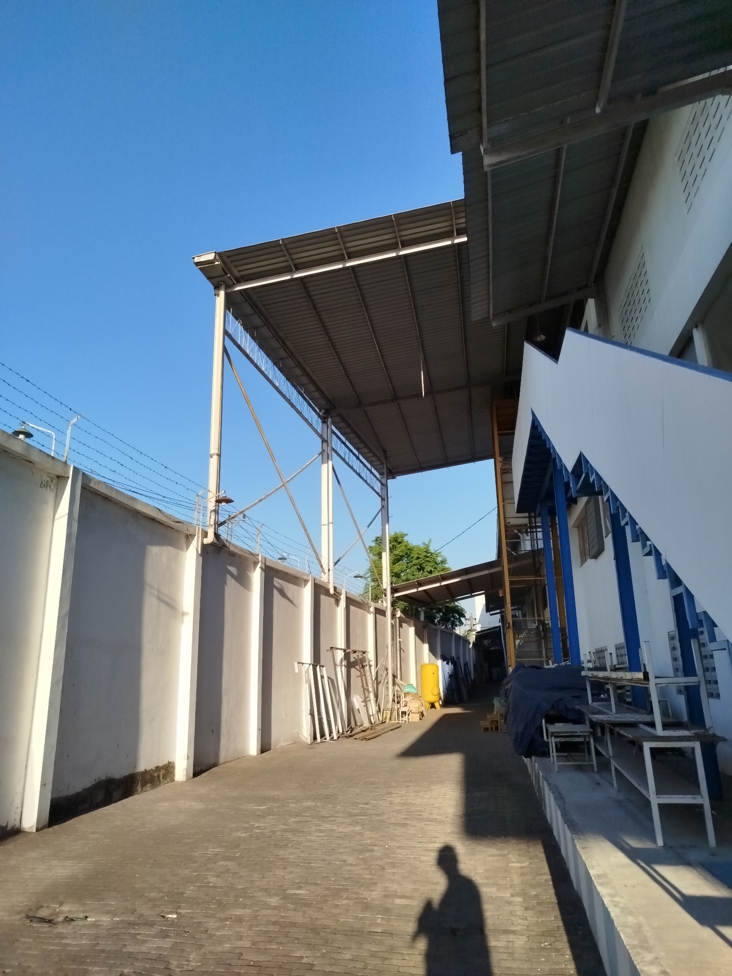 The image shows a concrete pathway lined by a tall, light-colored concrete wall with barbed wire on top on the left. A corrugated metal roof structure supported by metal poles extends over the pathway from the right. The wall casts long shadows across the pathway, which is paved with rectangular bricks.
To the right of the pathway, a white building with blue accents is partially visible. This building has a series of white metal shelves or benches in front of it. Further down the pathway, a yellow cylindrical tank is present, along with various metal frames and construction materials. The sky is clear and blue.
The shadow of a person holding a phone is visible on the pathway in the foreground. This scene is consistent with an industrial or storage area within the city of Surakarta, Indonesia, possibly a factory or warehouse yard.