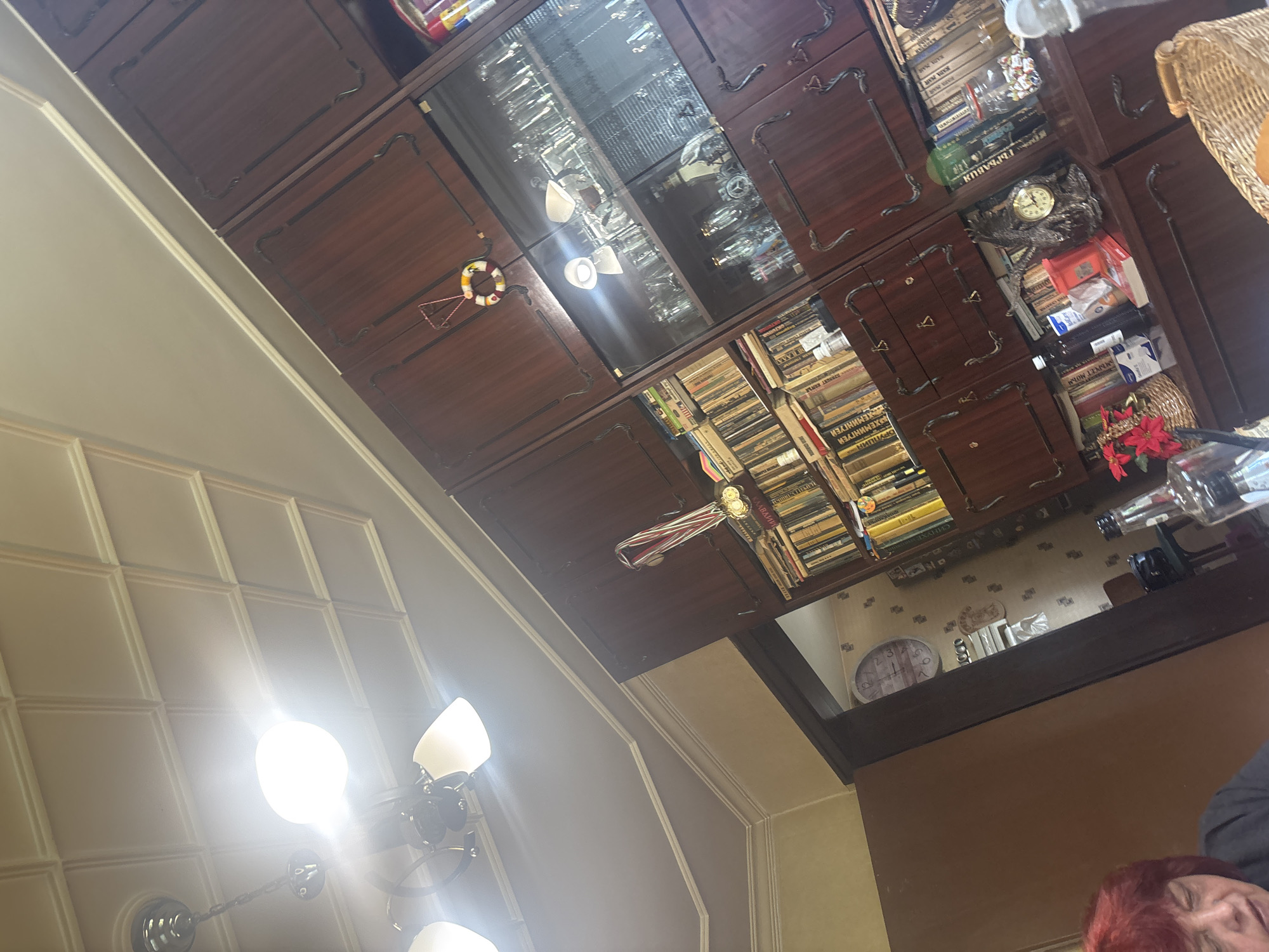 The image depicts an interior scene, likely a living room or study, in a residential setting. Dominating the frame is a large, dark wood built-in cabinet filled with books and glassware. The cabinet has ornate detailing and glass-fronted sections showcasing various items. On one shelf, a clock is visible, as are several bottles and containers. Further to the left, a doorway leads to another area where a round clock with Roman numerals is mounted on the wall. A portion of a woman with red hair is visible in the lower left of the frame, facing away from the camera and appearing to be seated. Above the cabinet, a modern chandelier with multiple lights illuminates the ceiling, which features a coffered design. The overall impression is one of a well-furnished, possibly older, home. The image was taken in Pernik, Bulgaria.