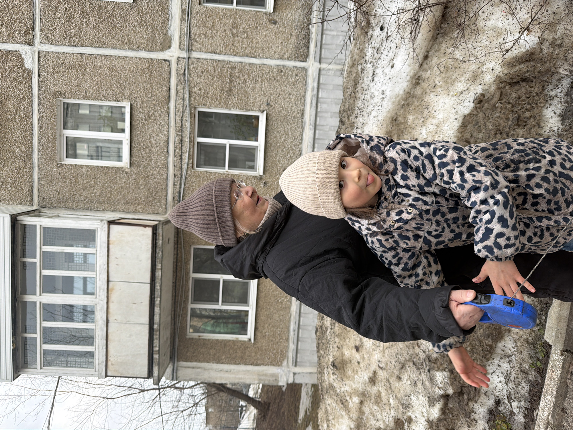 The scene depicts an adult female and a young female child outdoors in Perm, Russia. The adult, positioned partially behind the child, wears a brown ribbed knit beanie, round eyeglasses, a light-colored turtleneck, and a black puffy jacket. She holds a blue retractable leash in her right hand and her gaze is directed downward towards the child's left side. The child, standing in the foreground, wears a light beige ribbed knit beanie and a hooded leopard-print winter coat. She faces the viewer with a playful expression, her tongue extended. Her right hand is in contact with the adult's hand holding the leash. The background features a multi-story residential building with a textured, grey-brown pebbledash facade, distinct white-framed windows, and an enclosed balcony on the upper left. The ground is characterized by patches of melting, dirty snow interspersed with bare brown earth. Sparse, leafless tree branches are visible on the periphery of the frame, and a concrete curb runs along the lower foreground.