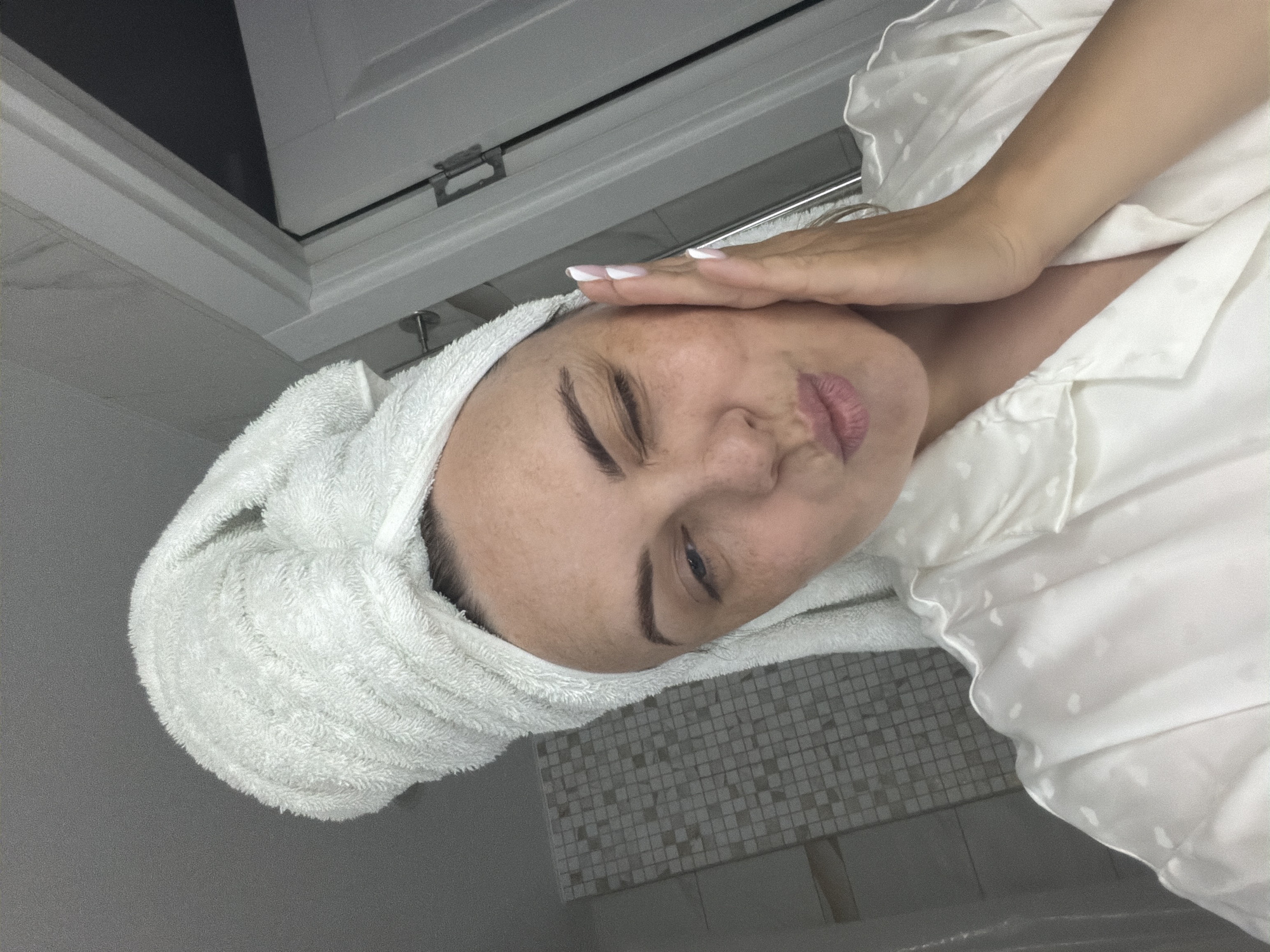 A woman with a white towel wrapped around her head stands in a bathroom. She is wearing a white satin pajama top and is touching her forehead with her left hand. Her fingernails are painted with a white tip French manicure. Her lips are puckered. The bathroom features white cabinets, gray and white tiled walls, and gray flooring. The location is in a private home, potentially in Votkinsk, Russia, but there is no information to independently verify this.