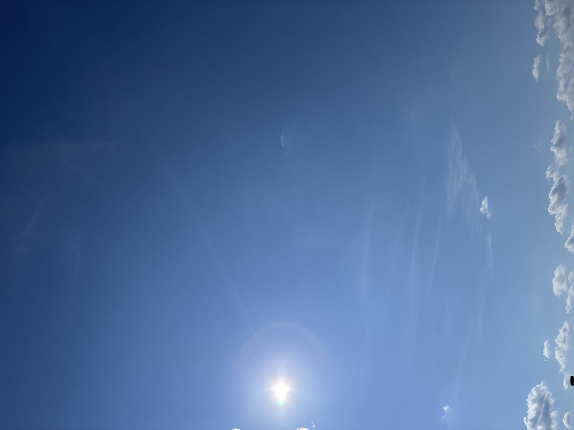 The image depicts a bright blue sky with a few scattered white clouds. The sun is visible in the upper left quadrant, casting a lens flare across the sky. The lower portion of the image shows a sliver of what appears to be a building or structure, with some white clouds gathered around it. There are no people visible in the image. The scene suggests an outdoor environment, possibly a rural area given the presence of open sky and clouds. The location in Ozyaritska-Slabadski rural council, Belarus, would imply a likely natural landscape.