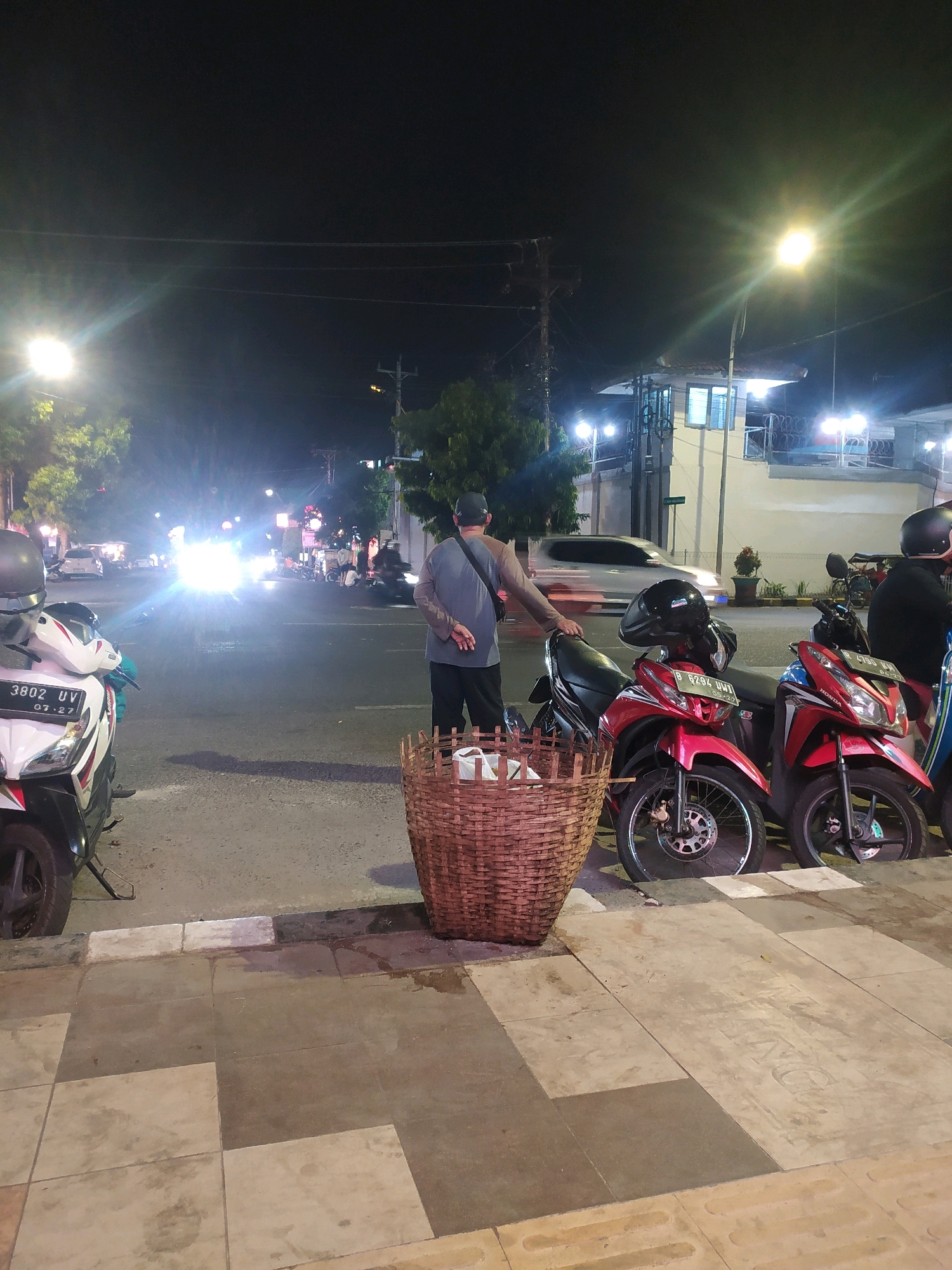 A nighttime street scene in Purbalingga, Indonesia, is depicted. In the foreground, a woven basket sits on a sidewalk next to a white and red motorcycle. Across the street, several red motorcycles are parked. A man in a gray shirt and dark pants stands near the basket, facing away from the camera and towards the street. The street is lit by streetlights and the headlights of vehicles, some of which are blurred due to motion. Buildings and trees are visible in the background.