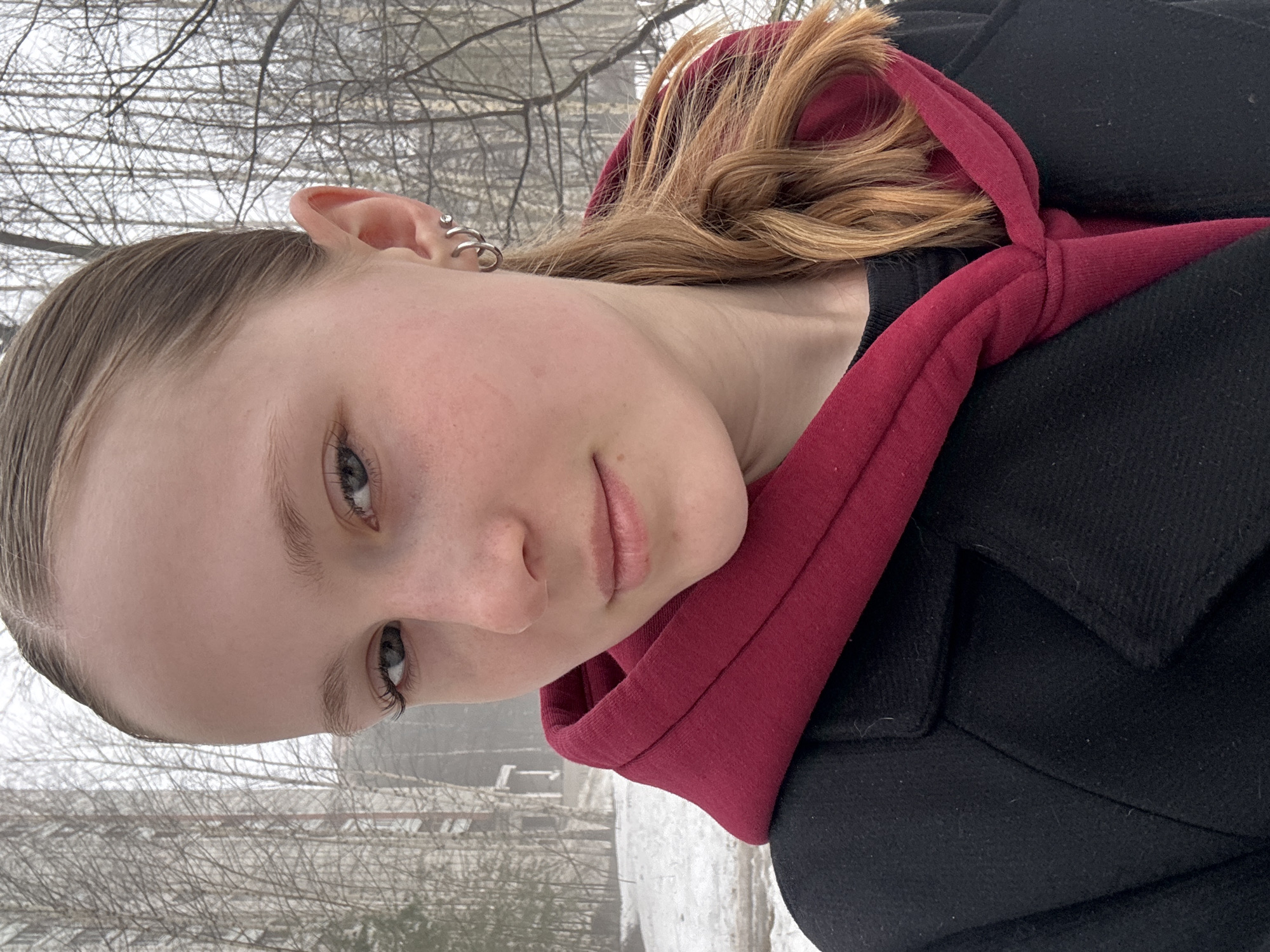 A young woman is pictured in an outdoor setting, likely during winter, given the bare trees and hints of snow on the ground. She is wearing a black coat with a maroon-colored hoodie underneath. She has light skin, light-colored hair pulled back at the front and loose in the back, and multiple piercings in her ear. The background appears to be a residential area with buildings visible behind the trees. The location is Chaikovsky, Russia.