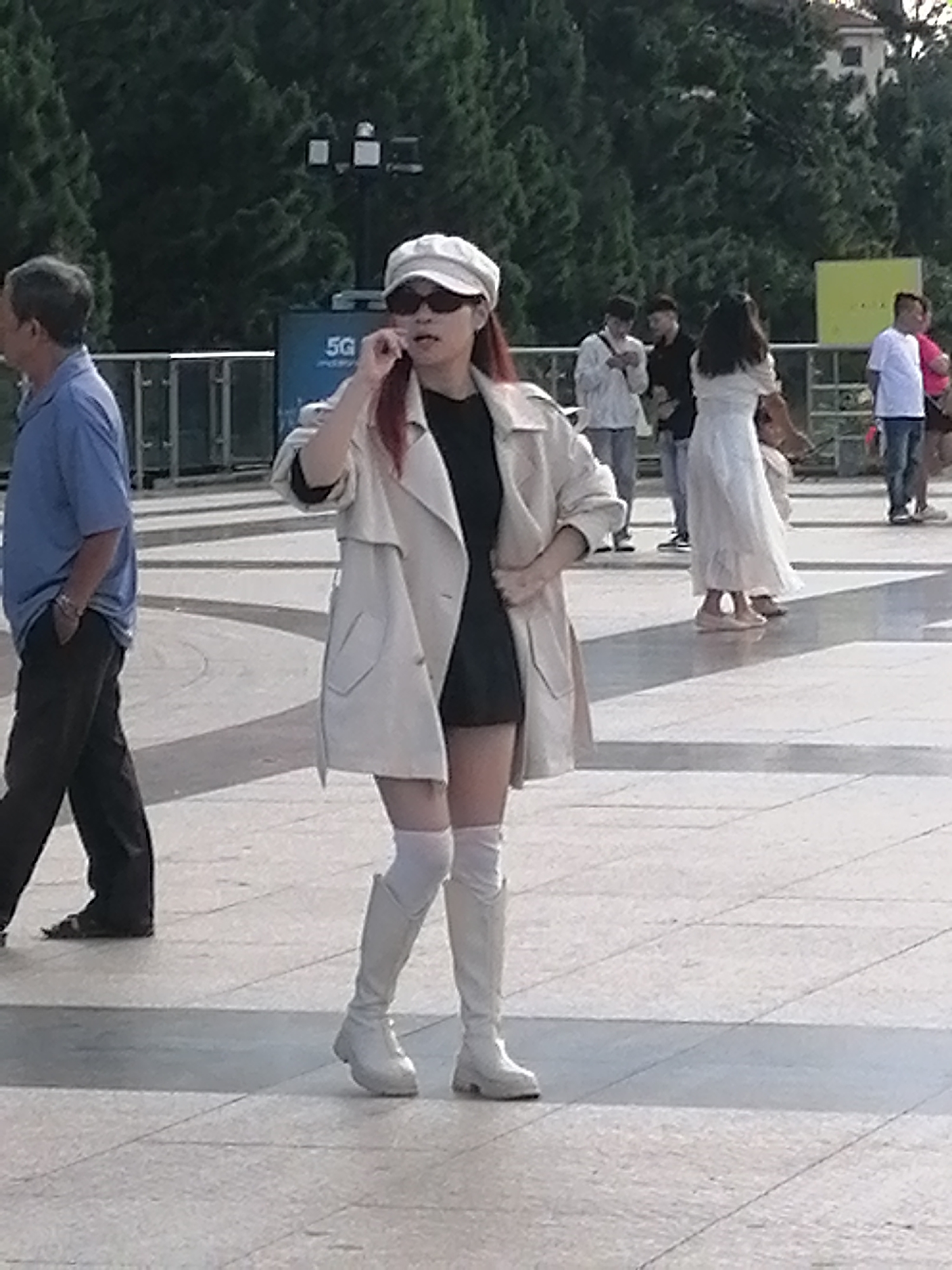 A woman in a light-colored trench coat, dark dress, white knee-high boots, and a white cap is standing on a paved plaza in Da Lat, Vietnam. She is holding a cell phone to her ear. Several other people are visible in the background, including a man in a blue shirt walking to the left, and a group of people standing near a fence. Trees line the background of the scene. The plaza is paved with large, light-colored tiles, and there are shadows cast across the ground.