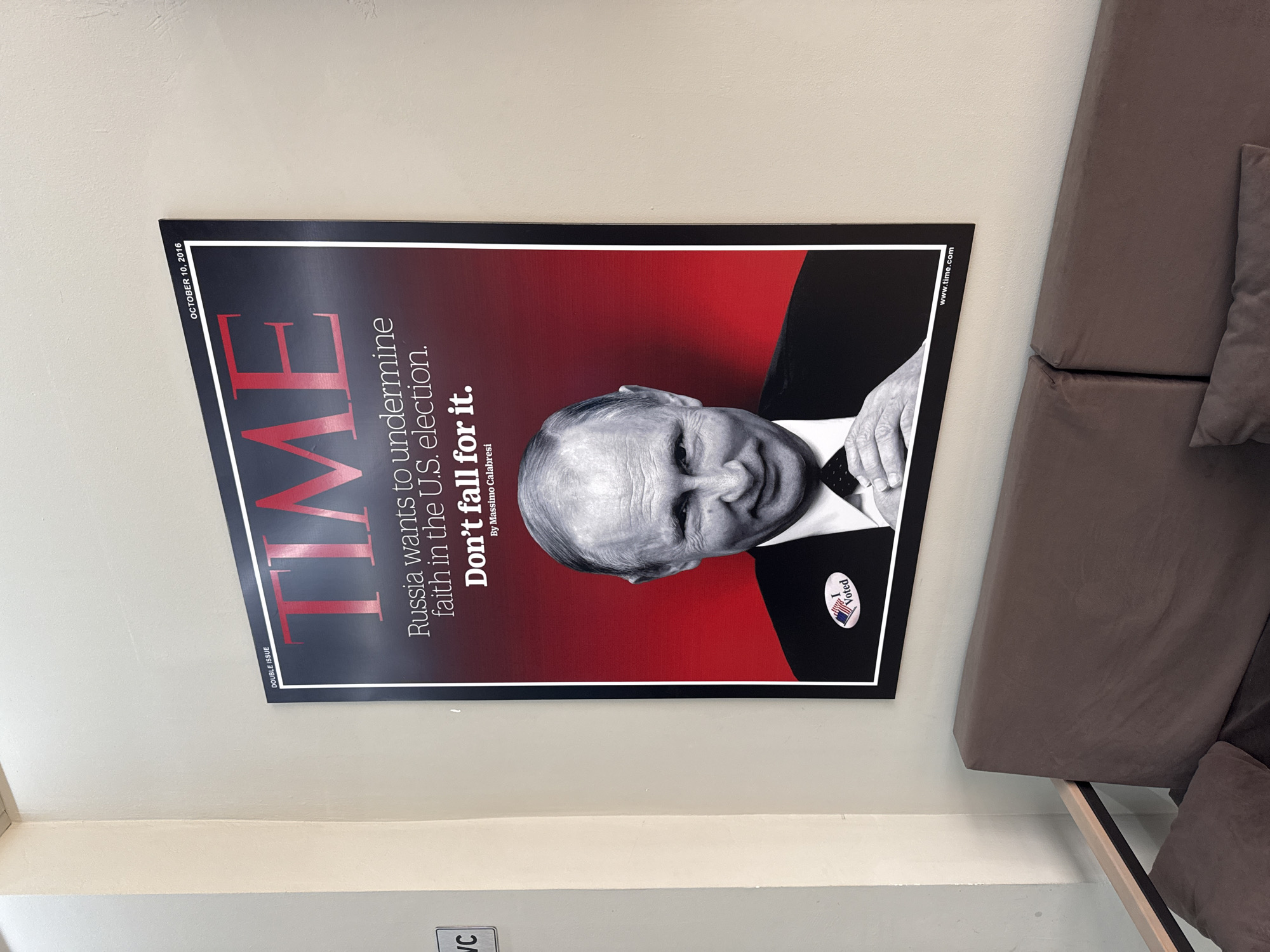 The image shows a framed Time magazine cover featuring Vladimir Putin. The cover has the headline "Russia wants to undermine faith in the U.S. election. Don't fall for it." Putin is wearing a suit and tie and has an "I Voted" sticker on his lapel. The magazine cover is hung on a beige wall. In the foreground, a brown sofa with pillows is visible. The scene appears to be an indoor space, possibly a cafe or a waiting area, and is not located in Chaikovsky, Russia.