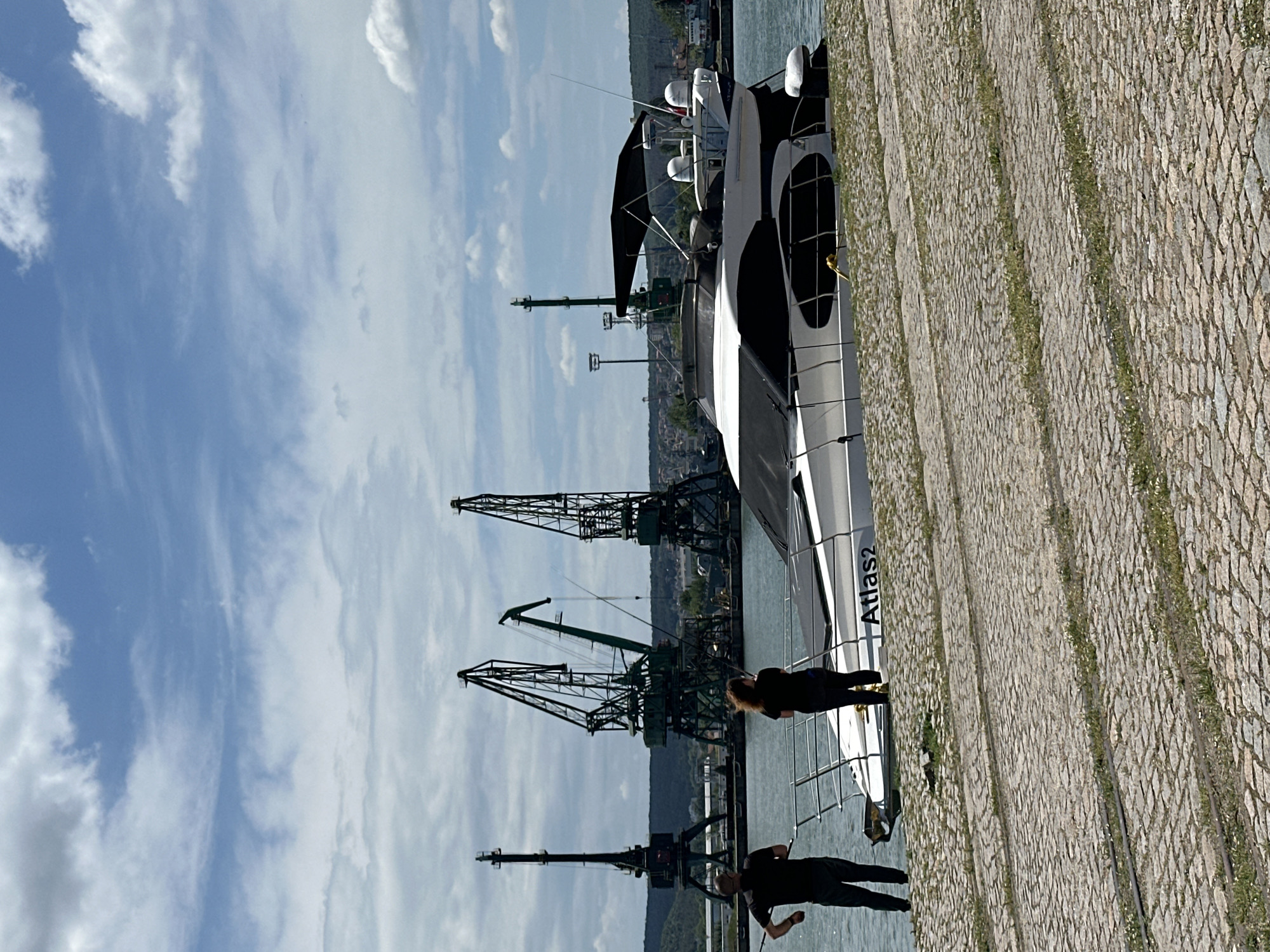 The image shows a harbor scene in Varna, Bulgaria, with several prominent cranes visible against a partly cloudy sky. A modern-looking yacht, named "Atlas2", is docked alongside a paved quay. Two individuals are present: one person is standing closer to the vessel, and another is walking along the quay. The waterfront is constructed with cobblestones, with small areas of vegetation growing between them. The city's buildings are visible in the background.