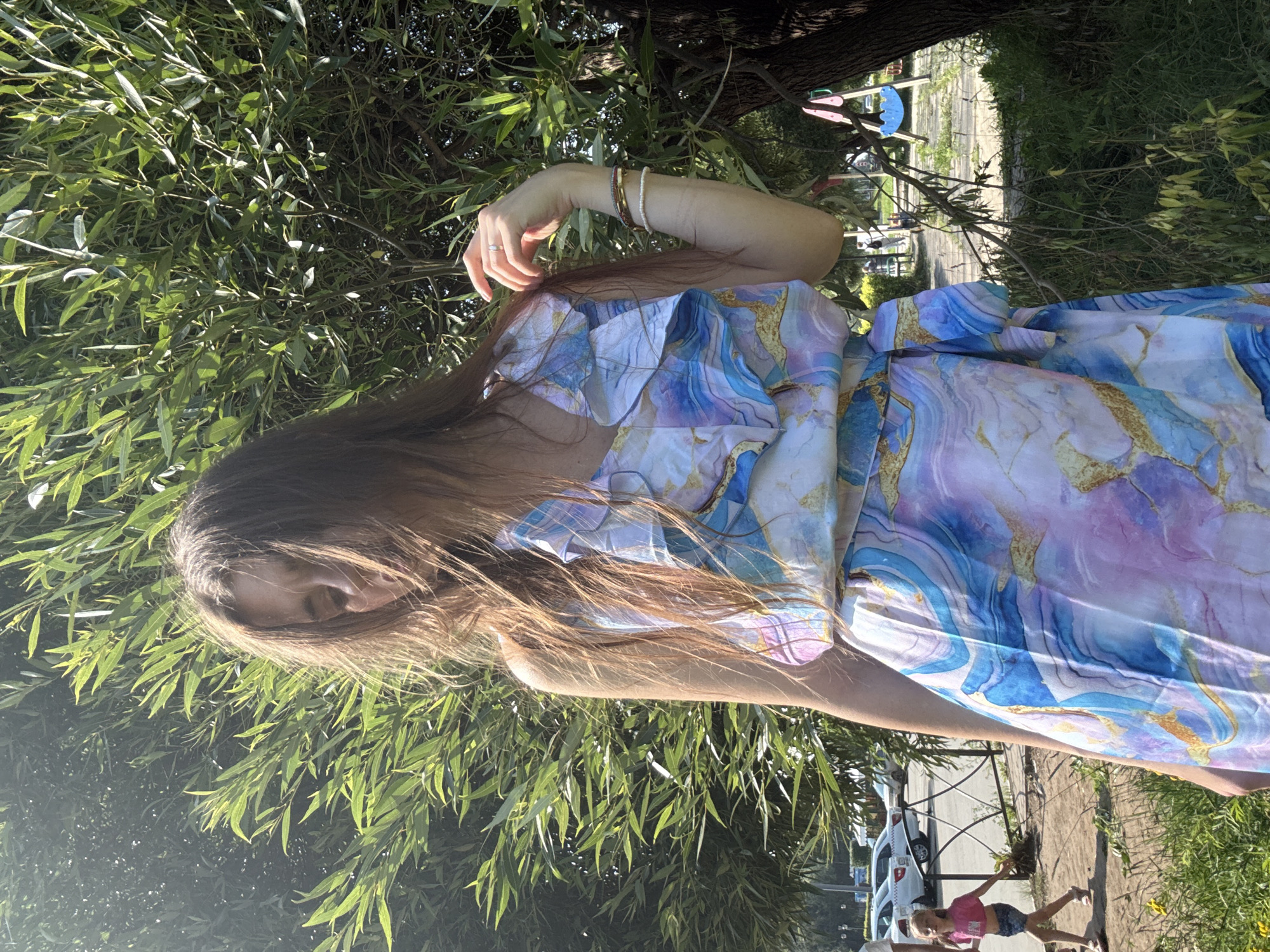 In the city of Unknown, Unknown, a young woman with long, brown hair stands outdoors, her face partially obscured by her hair and the bright sunlight. She is wearing a flowing dress with a colorful, marbled pattern of blues, purples, and gold. The dress has ruffled detailing around the neckline and a cinched waist with a tie belt. She is adorned with several bracelets on her right wrist and a ring on her right ring finger. Her right hand is raised, touching her hair near her face.
Behind her, a large tree with dense green foliage dominates the background, with sunlight filtering through the leaves. To the left of the woman, a young girl in a pink top and denim shorts walks away from the camera. Beyond the girl, a street with parked cars is visible, bordered by a metal fence. In the distance, a playground area with swings can be seen. The ground in the foreground is covered with grass and some yellow flowers. The scene appears to be a sunny day in a park or a public outdoor space.