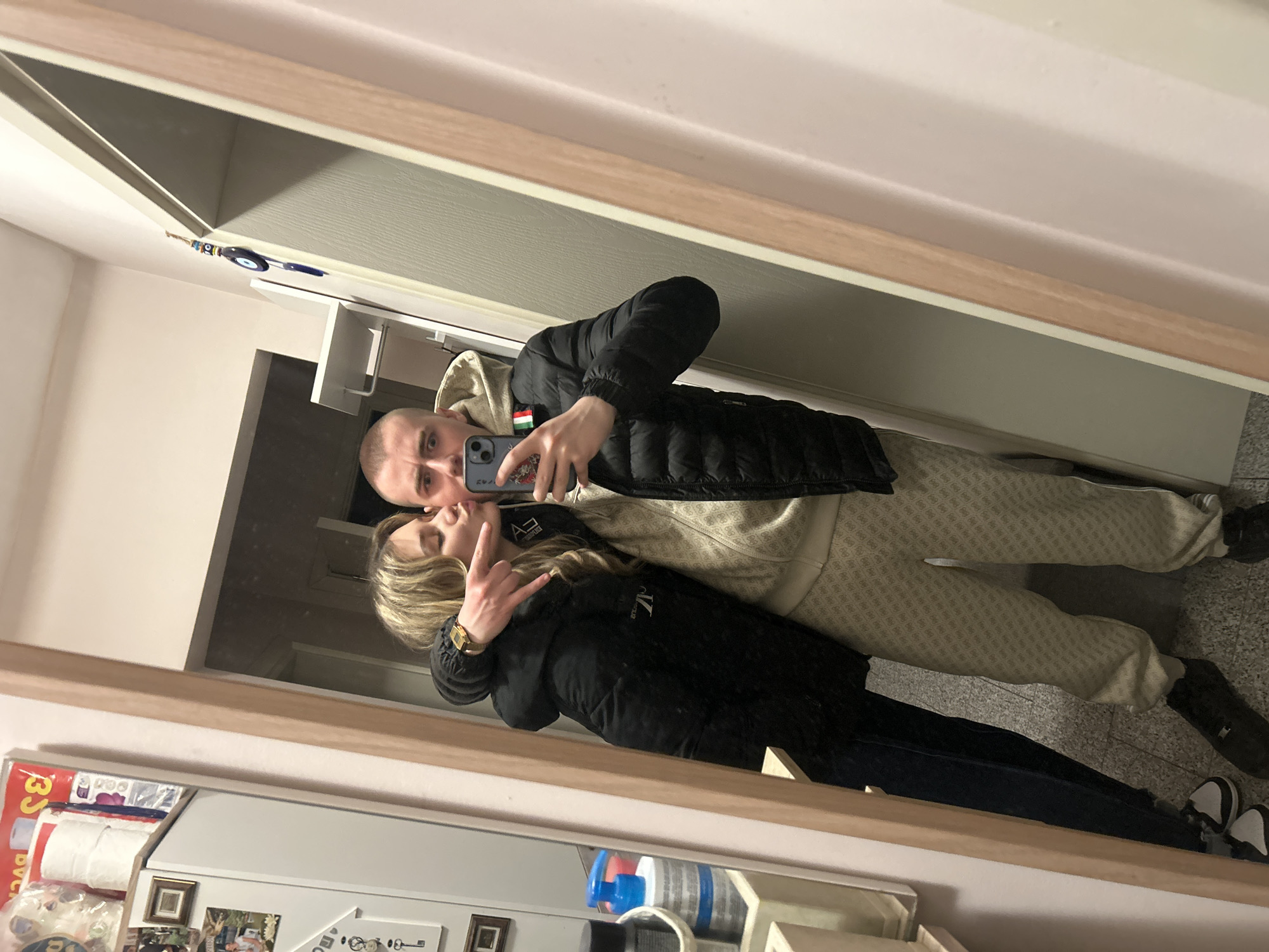 A man and woman are taking a selfie in a mirror. The man has a shaved head and is wearing a black puffer jacket and a hooded sweatshirt. The woman is wearing a black jacket and a gold watch. The background includes a refrigerator with pictures and magnets on it, a door, and a hallway. The scene is likely inside an apartment or house. There is an evil eye charm hanging above the door. The location cannot be determined as Sofia, Bulgaria.
