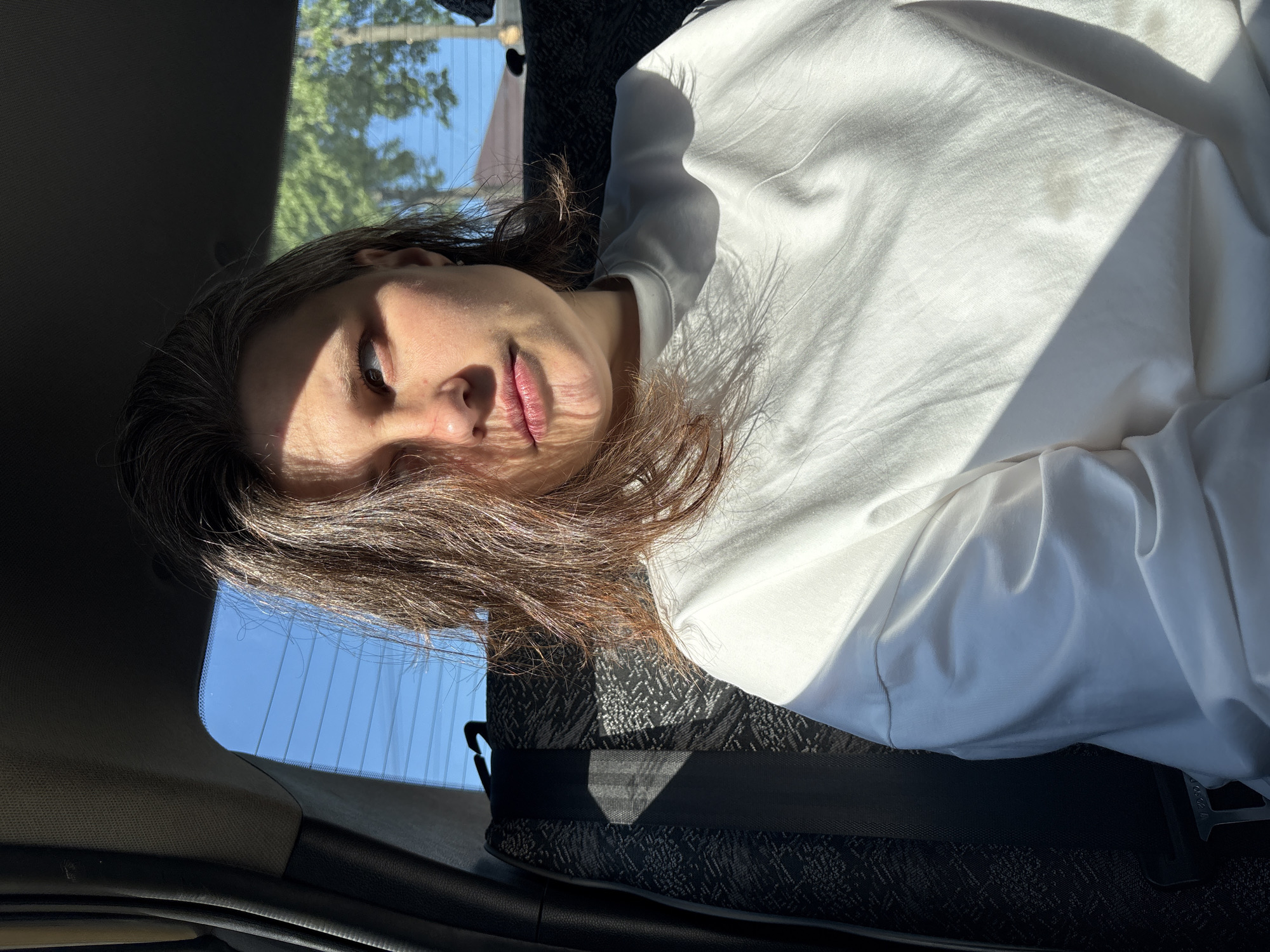 A person is lying in the backseat of a car. The person has light brown hair and is wearing a white sweatshirt. Sunlight is streaming through the car window, casting shadows on the person's face and clothing. The car interior is black and gray. Outside the window, trees and a blue sky are visible. There is no indication of the location in the city of Chaikovsky, Russia in this image.