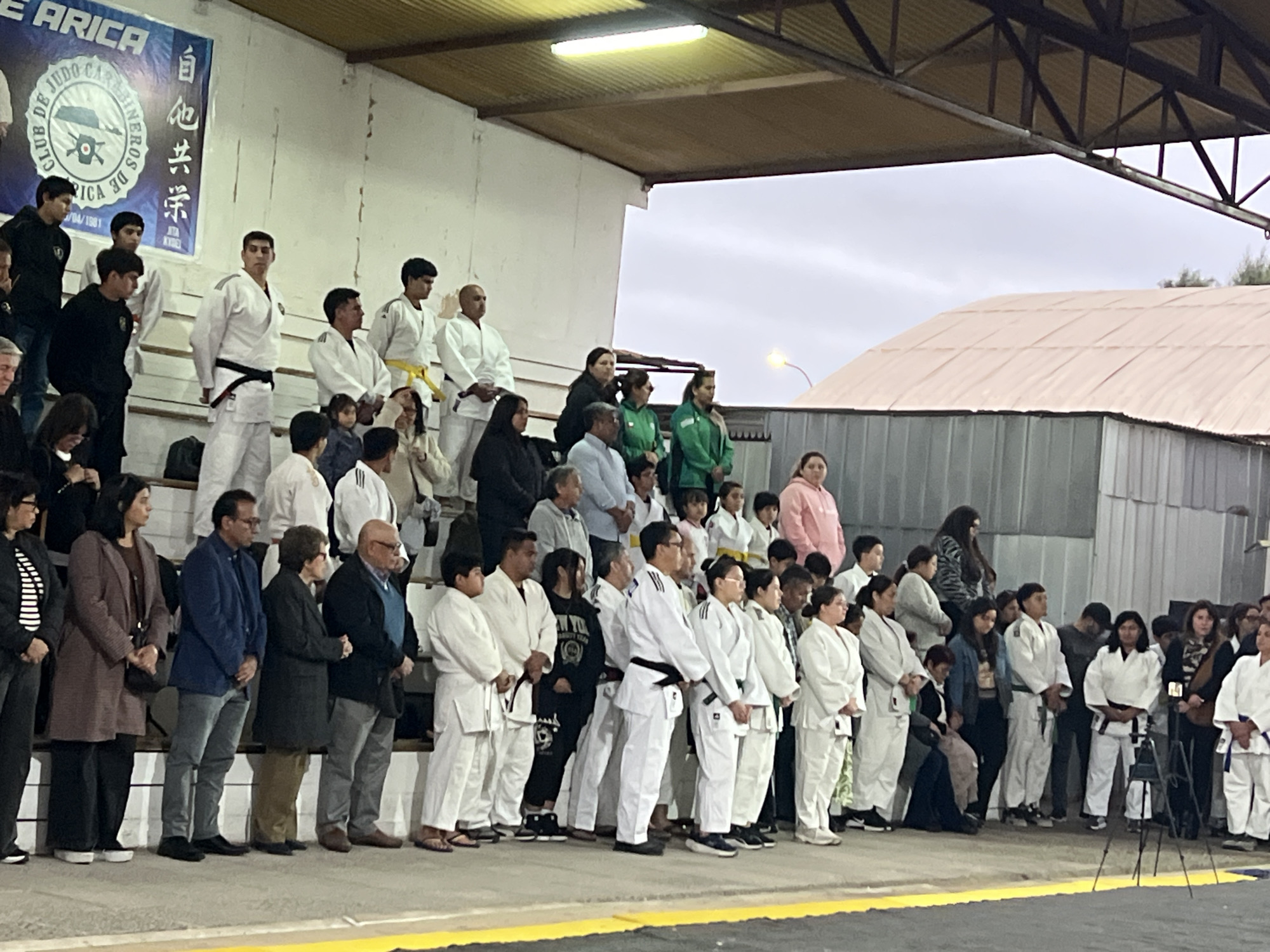 The image shows a group of people, predominantly young individuals in judo uniforms, gathered on a raised platform under an awning. The judo uniforms are white, with some participants wearing colored belts. Several adults are also present, some in civilian clothing and others also in judo attire. The backdrop features a white wall with a blue banner that has text and graphics, identifying the event as originating from "CLUB DE JUDO CARABINEROS DE ARICA". This banner indicates the location is in Arica, Chile, and the event is related to judo. To the right of the platform, a corrugated metal structure with a pink roof is visible. The sky above is overcast. The scene appears to be an indoor or semi-outdoor event, possibly a competition or a ceremony, given the formal attire of many participants and the banner.