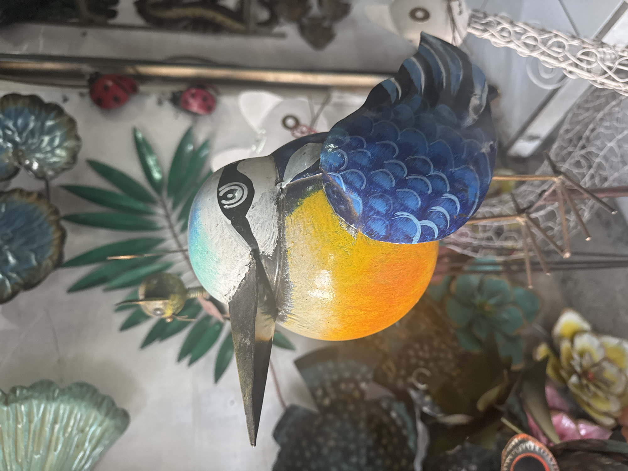 A close-up shot shows a painted, wooden bird figurine with a pointed beak and blue and orange plumage. It is partially visible in a cluttered, artificial environment. Two small red ladybug decorations sit on a shelf near leafy green and teal metal artwork. Above, a white, decorative plastic chain hangs with other indistinct objects. The photo was taken in the Ubud District of Indonesia.