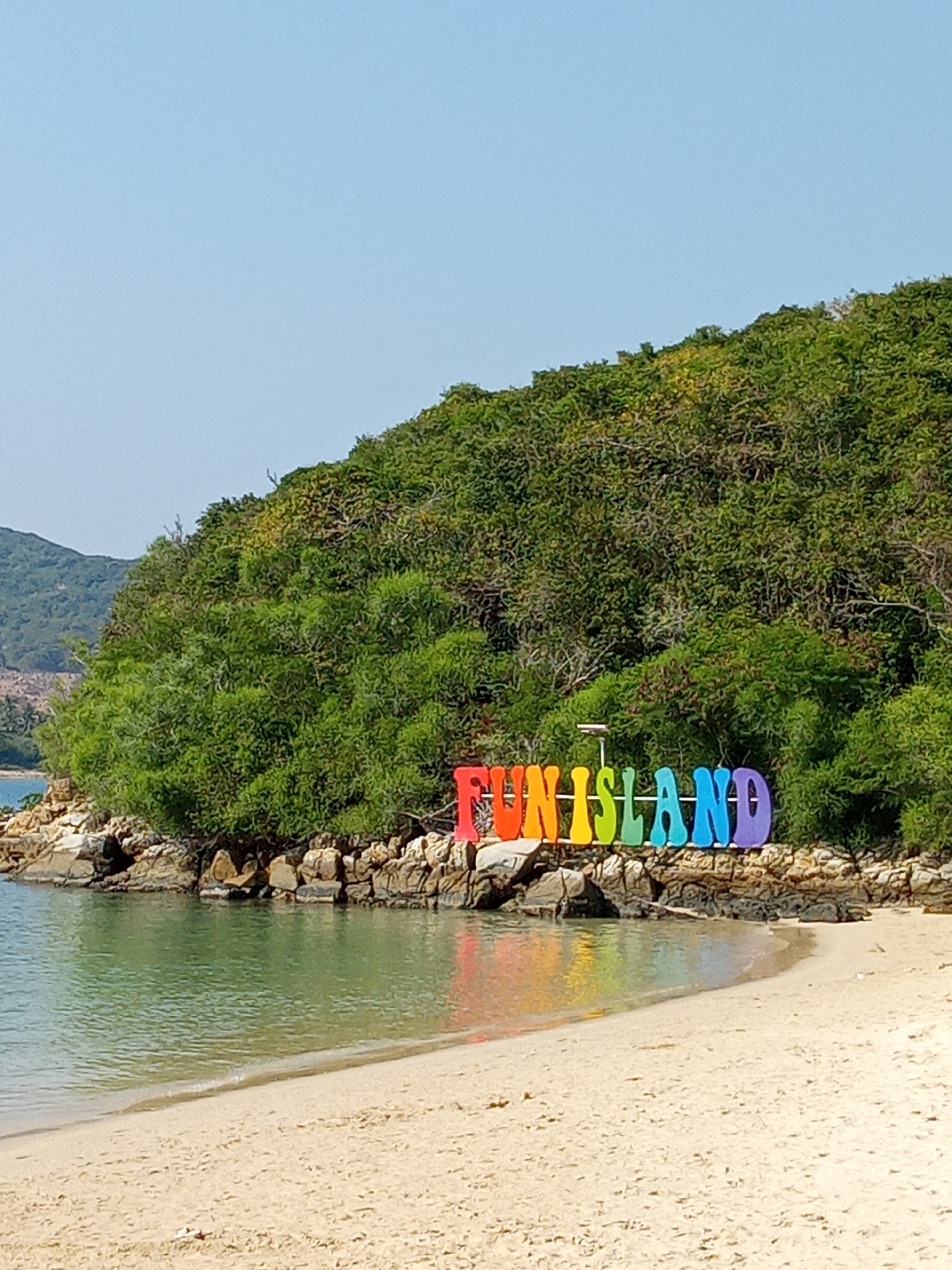 The photograph shows a beach in Nha Trang, Vietnam. The shoreline has light-colored sand meeting turquoise water. Behind the beach, lush green vegetation covers a hillside. A large, colorful sign that reads "FUN-ISLAND" is placed among the rocks at the base of the hill, with the letters in rainbow colors. The sign's reflection is visible in the water. The sky is clear and light blue. A distant mountain is visible on the left.