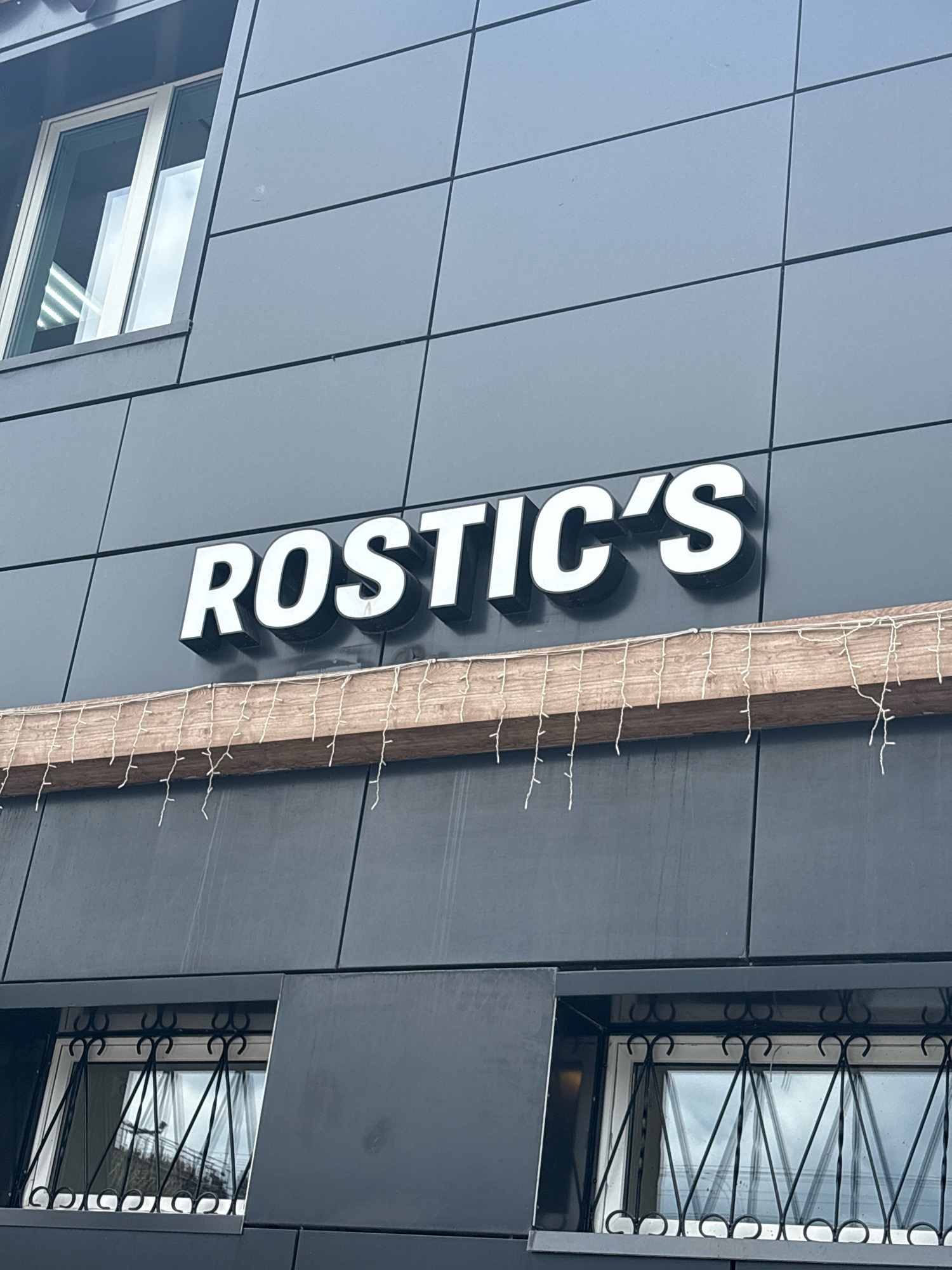 A photograph captures the exterior signage of a business named "ROSTIC'S." The white, three-dimensional letters are mounted on a dark gray, tiled building facade. Below the signage, a rustic wooden beam is attached, adorned with a string of small, white lights that hang down like icicles. The building has several windows, one of which is visible in the upper left corner and appears to be a modern window with a light-colored frame. The lower section of the building features two windows with decorative black metal grilles. The overall impression is of a commercial establishment with a somewhat modern yet rustic aesthetic. The location is specified as Unknown, Unknown.