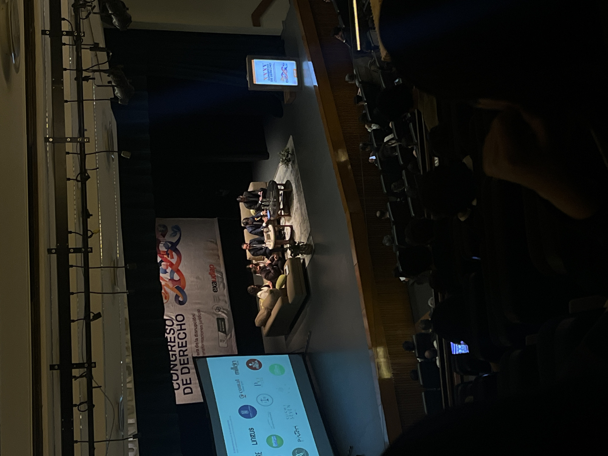 The image shows a view from the audience of a stage in what appears to be a conference hall in San Andrés Cholula, Mexico. The stage has a panel of people seated on sofas and chairs. There's a screen on the left with sponsor logos and a backdrop with "CONGRESO DE DERECHO" (Law Congress) written on it. Another screen displays a similar message, possibly the event's name. The audience is seated in rows of chairs, with some people using their phones. Lighting fixtures are visible on the left.