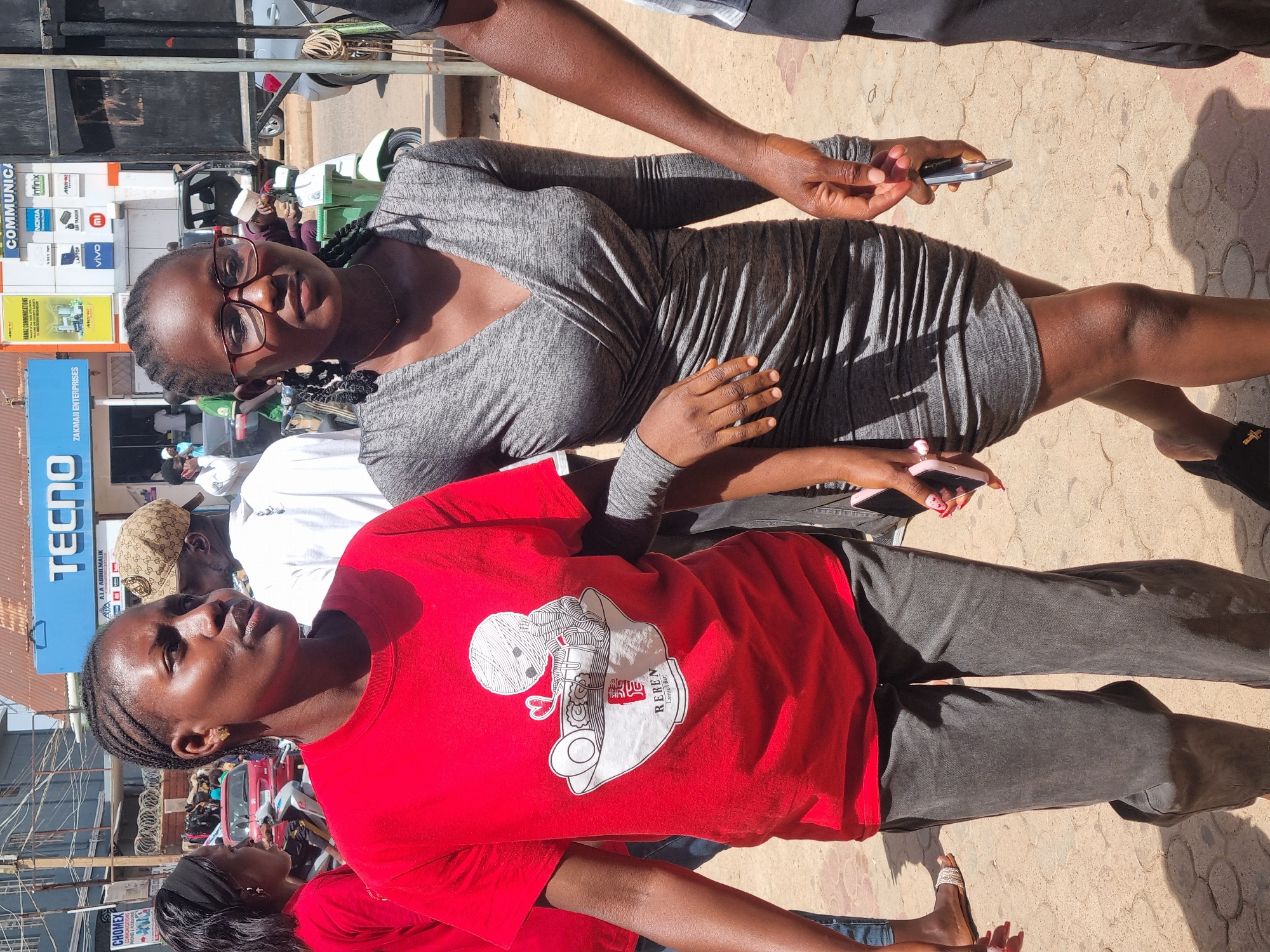 Two young women stand on a paved sidewalk in the city of Jos, Nigeria. The woman in the foreground wears a red t-shirt with a white graphic featuring noodles. She has braided hair and grey pants. The woman behind her wears a grey dress and glasses. Both women hold smartphones. A man’s arm is visible above the woman in the grey dress. Storefronts are visible in the background, displaying brands like Tecno, Infinix, and Nokia. Signage includes “ZAKMAN ENTERPRISES”, “AAALA ABDULMALIK”, and “HANAZ COMMUNICATIONS”. Motorcycles and other vehicles are parked on the street.