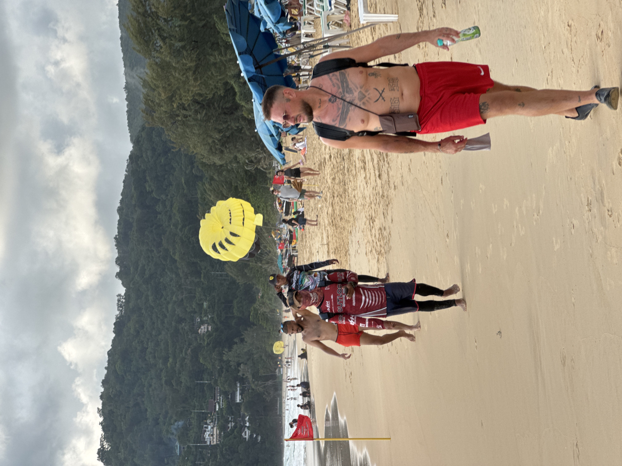 The image captures a scene on Karon Beach in Karon, Thailand. The beach is sandy and stretches towards a lush, tree-covered hillside in the background. The sky is overcast with scattered clouds.
In the foreground, a man with numerous tattoos on his chest and legs is walking towards the viewer. He is wearing red shorts, sunglasses, and a small brown satchel over his shoulder. He is holding a bottle of water in his right hand. Behind him, a group of three men are walking away from the viewer. The man in the middle has a shirtless torso and is wearing red shorts. The man to his left is wearing a dark long-sleeved shirt with colorful graphics and dark shorts. The man furthest to the left is wearing a red and white long-sleeved shirt and dark shorts with red trim, and is carrying a phone in his hand.
Further back on the beach, there are several other people walking and sitting. A bright yellow parasail is visible in the sky above the beach, suggesting parasailing activities. The shoreline is visible with gentle waves. Beach umbrellas and lounge chairs are set up further back on the sand, with more people scattered around. A red flag on a yellow pole stands near the water's edge. The overall scene depicts a typical beach day with recreational activities.