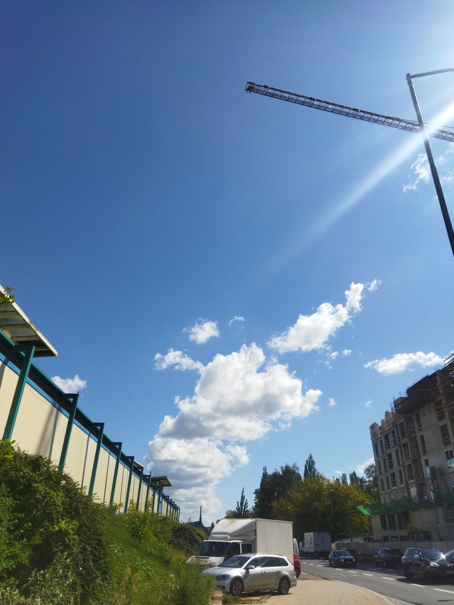 This is a daytime, medium-shot, eye-level photograph taken in Szczecin, Poland. The image captures a scene on a bright, sunny day with a clear blue sky and scattered white clouds.
In the foreground, to the left, is a grassy embankment covered with dense green foliage, leading up to a light-colored wall with a green trim. The wall appears to be part of a structure with a pitched roof. Further along the embankment, and to the right, are parked vehicles. These include a white panel van, a silver hatchback, and other cars driving on a road.
Dominating the upper portion of the image is a construction crane with its jib extended horizontally across the frame. To the right of the crane is a tall, modern streetlamp. In the background, on the right side of the image, is a multi-story building under construction, with visible scaffolding and concrete framework. Trees and vegetation are visible behind the construction site and lining the road.
The overall impression is that of an urban environment undergoing development on a pleasant, clear day.