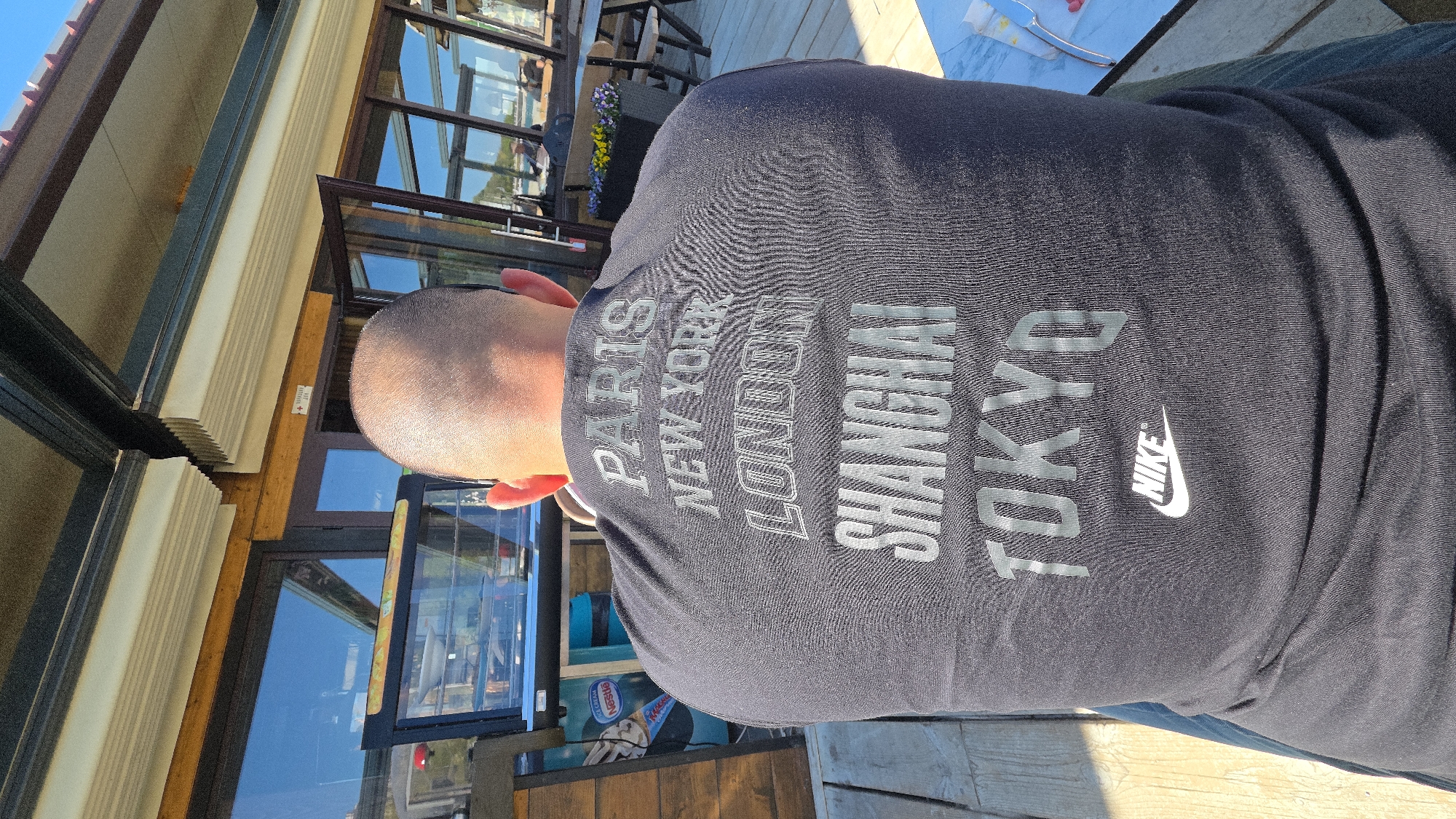 An eye-level shot of a man from behind, sitting at an outdoor venue in Varna, Bulgaria, wearing a dark blue t-shirt featuring the words "PARIS," "NEW YORK," "LONDON," "SHANGHAI," and "TOKYO" stacked vertically on the back, along with the Nike logo. The location appears to be a restaurant or cafe with outdoor seating. The background shows glass doors and windows with reflections of the surrounding environment, including what appears to be another building and some greenery. Tables and chairs are visible through the reflections. The man has a shaved head. The surface he is sitting on appears to be wooden planks. Part of another person's body, possibly jeans, is visible on the right edge of the frame.
