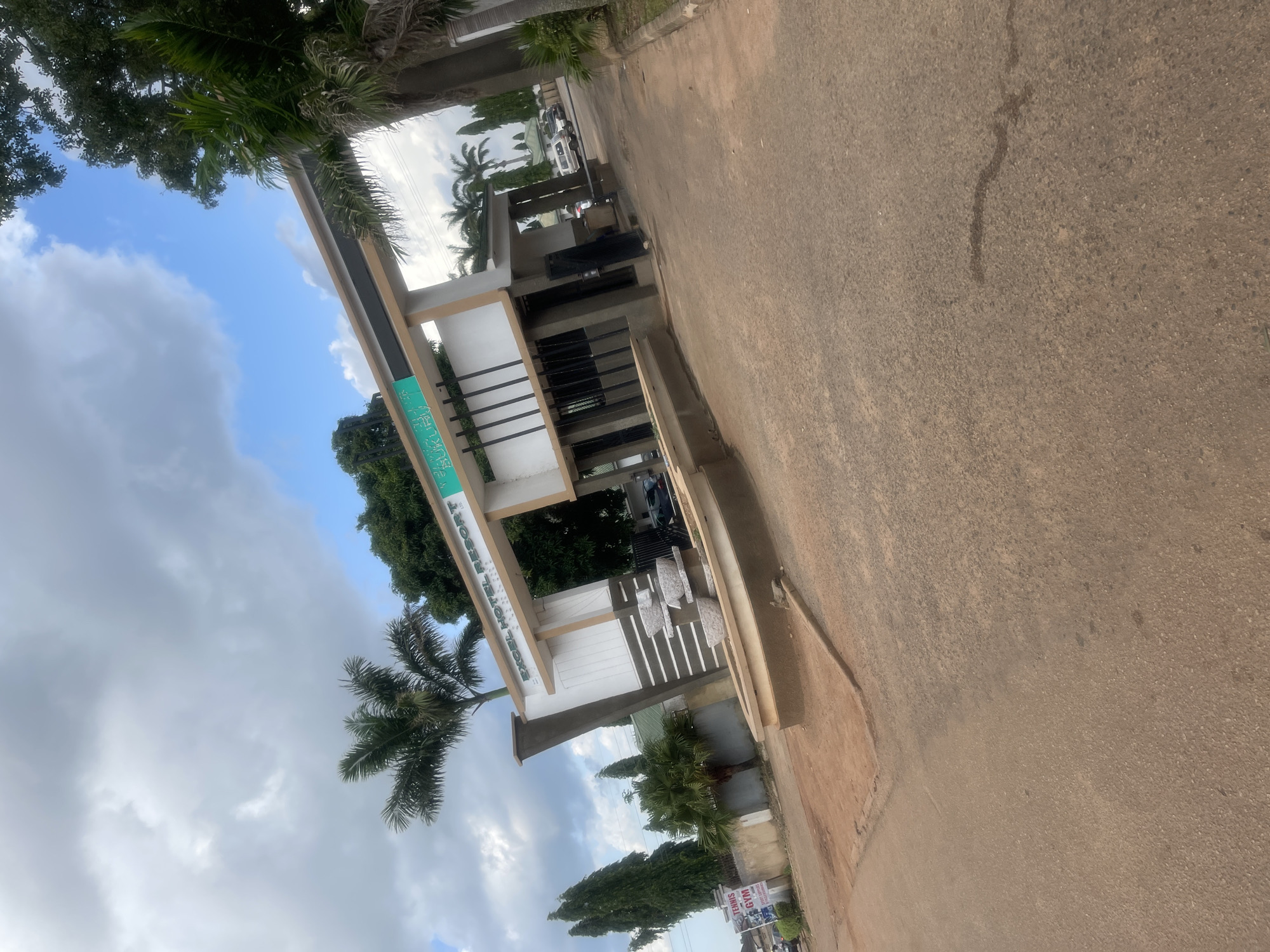 A gated entrance to "EXCEL HOTEL RESORT" in Bukuru, Nigeria, is visible under a partly cloudy sky. The resort entrance features a beige and white architectural design with a prominent archway. A sign displaying "EXCEL HOTEL RESORT" is mounted on the arch. To the left of the entrance, a sign for "TENNIS GYM SPORT COMPLEX" is posted on a pole. Several vehicles are parked within the premises. The surrounding environment includes palm trees and other vegetation, with a paved road leading into the resort and a dirt area to the left of the entrance. The ground in the foreground is a rough asphalt surface.