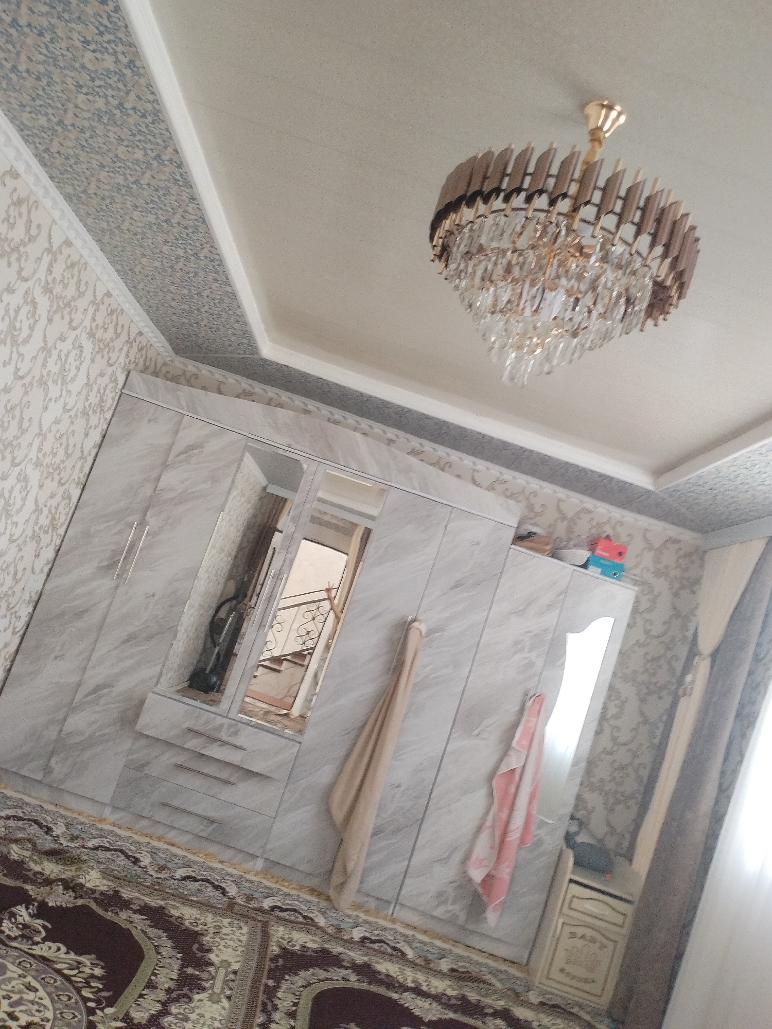 The image shows a bedroom in Kyrgyzstan. The room features a large, marbled wardrobe with mirrored doors. A chandelier with crystal accents hangs from the ceiling, which is decorated with patterned wallpaper and a geometric border. A patterned rug covers the floor. A towel hangs from the wardrobe, and a small chest of drawers is visible on the right side of the room. The scene is indoors, with natural light coming from a window on the right, suggesting it is daytime. The overall impression is of a well-decorated and tidy room. There is no visible text in the image.