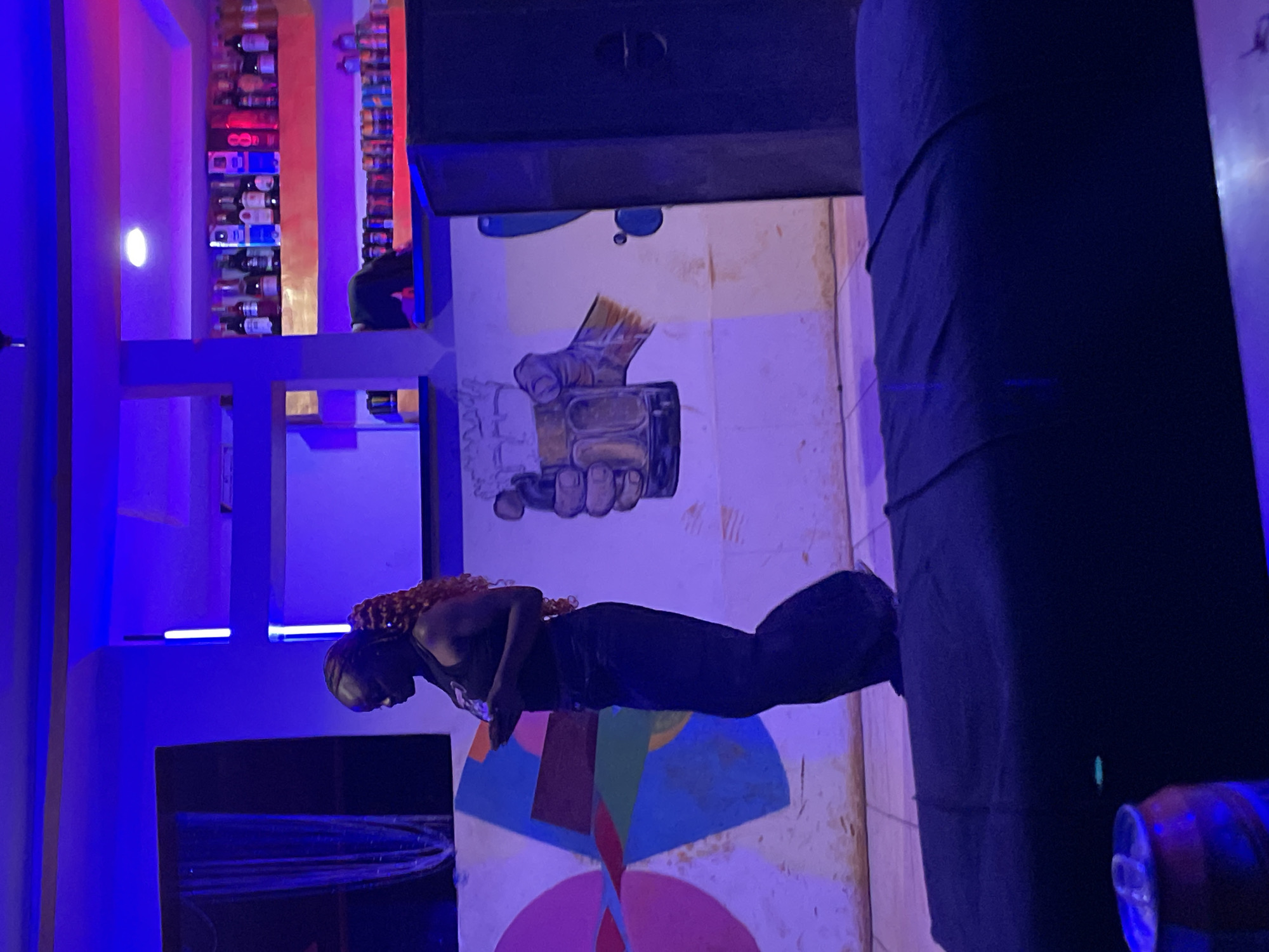 In an indoor setting, possibly a bar or lounge in Durumi, Nigeria, a person with curly reddish-brown hair is seen lying down, facing left. They are wearing a dark tank top and dark pants that appear to be flowing or gathered around their legs, suggesting a tail-like shape. The person is positioned on a colorful, circular mural painted on the floor, which features segments of various colors like blue, pink, red, green, and yellow.
Behind the person, a mural is painted on the wall depicting a hand holding a glass with liquid and ice. The overall scene is dimly lit and bathed in vibrant blue and pink neon lights, typical of a nightlife venue. In the background, shelves stocked with numerous bottles of alcohol are visible, illuminated by the colored lighting. To the right, a large, dark, cylindrical object wrapped in fabric or material is prominent, partially obscuring the view of the wall and a person seated behind it.
The mood seems relaxed and perhaps artistic, given the murals and the person's pose. The time of day is likely evening or night due to the artificial lighting. No visible text is present in the image.