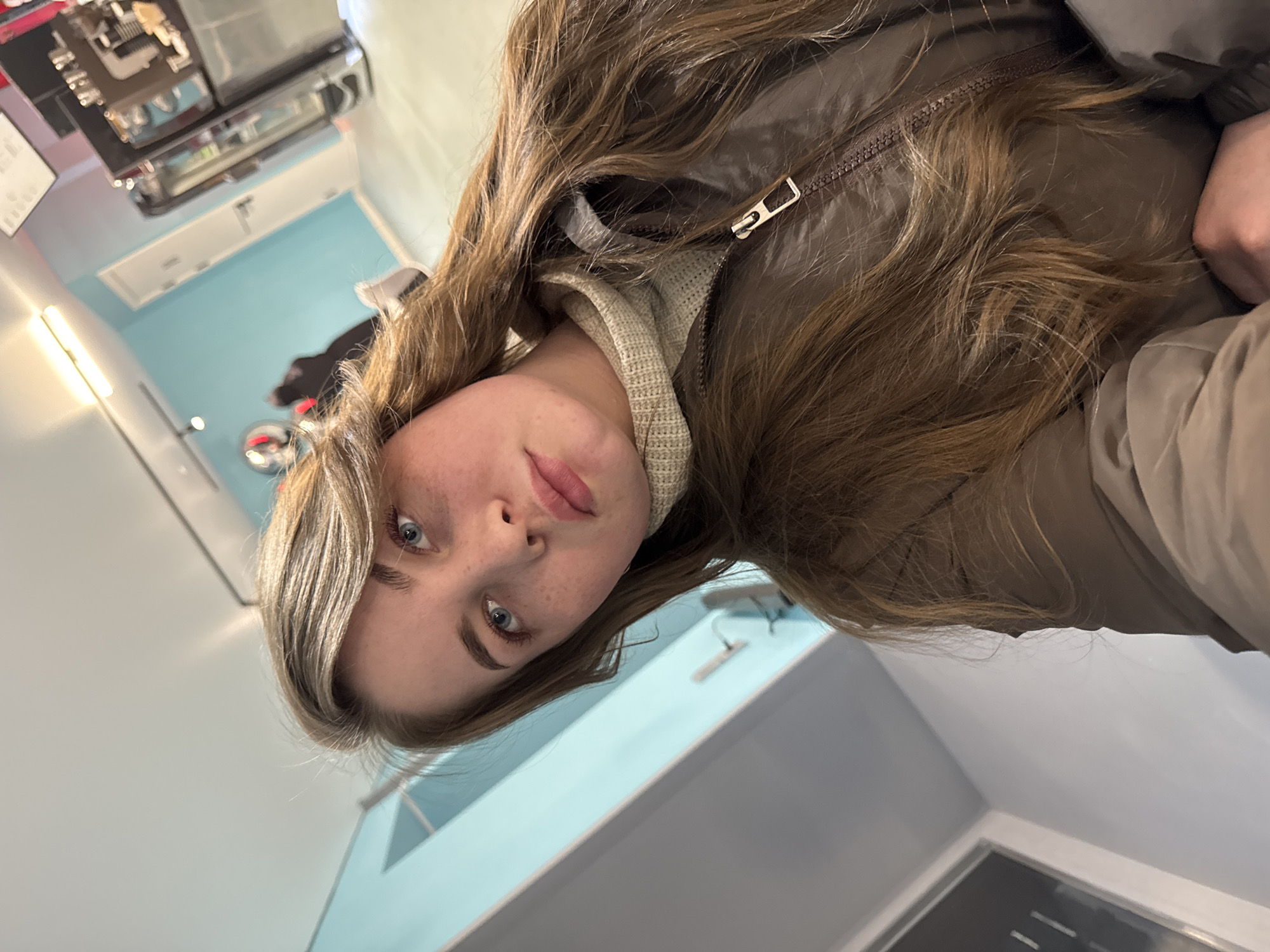 A close-up, slightly angled portrait features a young woman looking directly at the camera. She has long, wavy brown hair, blue eyes, and noticeable freckles across her nose and cheeks. Her lips are slightly parted, conveying a neutral to contemplative expression. She is dressed in a brown or olive-green puffer jacket with a metallic zipper, worn over a light-colored, possibly cream or beige, knitted turtleneck or high-collar top.
The setting is indoors, with light blue and grey walls visible in the background. Blurry elements behind her suggest a public indoor space, possibly a cafe, store, or waiting area, featuring a counter or display unit with reflections and artificial lighting. A circular object with a red center is also faintly visible in the background. The lighting is consistent with an indoor environment. There is no visible text in the image. While the location context of "Chaikovsky, Russia" is provided, there are no specific visual cues within the image itself to confirm this.