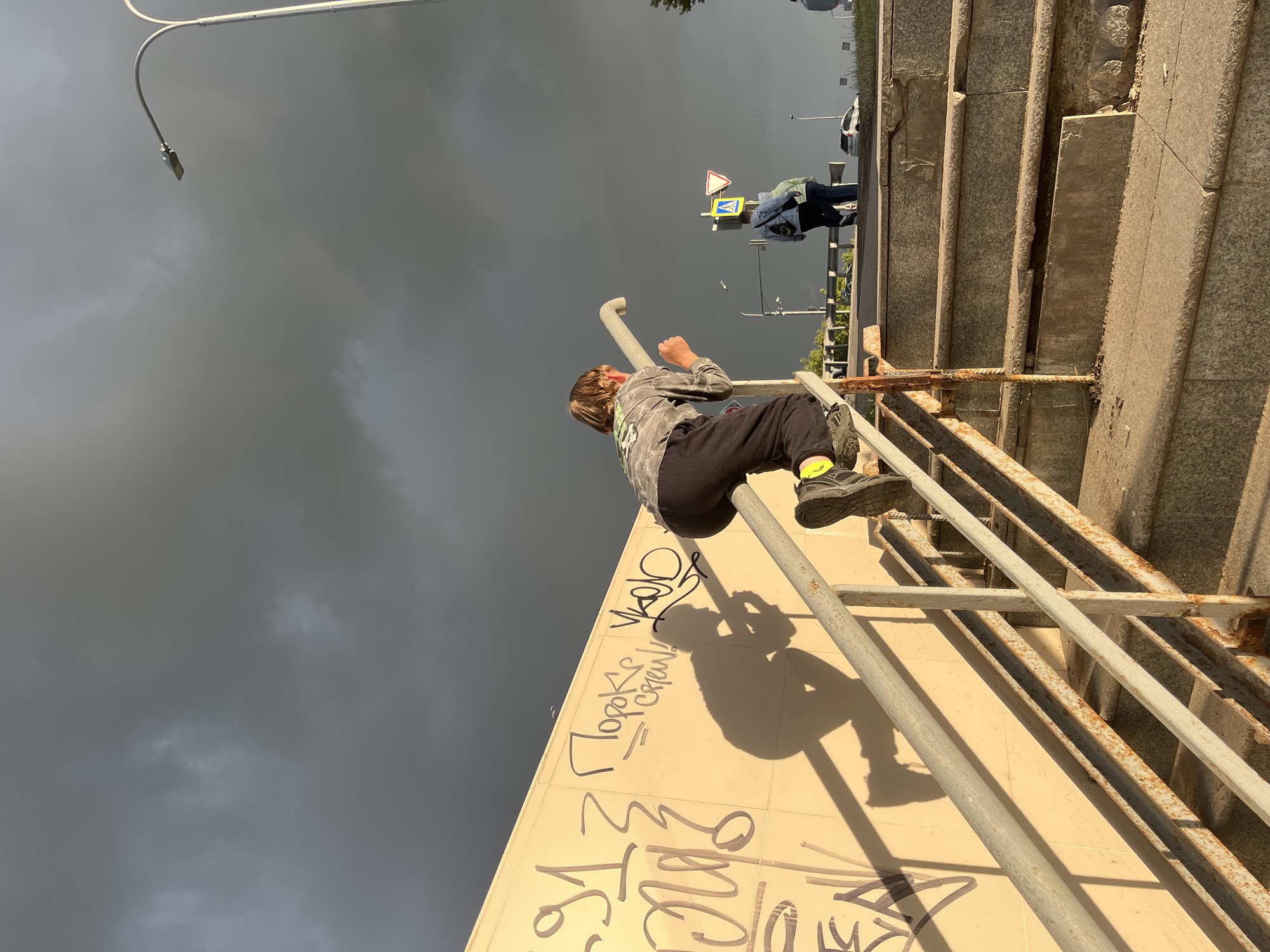 A young boy with light brown hair, wearing a grey and black patterned long-sleeved shirt and black pants, is captured in a dynamic pose as he climbs on a metal handrail. His left hand is firmly gripping the rail, and his right fist is clenched, suggesting exertion or determination. He is wearing black shoes with bright yellow accents.
The scene appears to be outdoors, possibly near a public space with stairs and a walkway. A set of stone stairs with metal railings is visible to the right of the boy. The background is dominated by a dark, cloudy sky, indicating overcast or potentially stormy weather.
In the distance, two individuals are walking on a paved area. One person is wearing a blue jacket and carrying a backpack, while the other is wearing a dark jacket. Traffic signs are also visible in the background, including a yellow pedestrian crossing sign and a triangular warning sign.
The wall below the handrail is covered in graffiti, with large black lettering clearly visible. One prominent phrase written in Cyrillic appears to be "Пороке!" which translates to "Vice!".
The lighting suggests it might be daytime, given the presence of shadows cast by the boy and the handrail, but the dark clouds create a dramatic and somewhat somber atmosphere. The overall impression is one of urban exploration or playful activity in a somewhat gritty environment. The location context provided is Nizhny Novgorod, Russia.