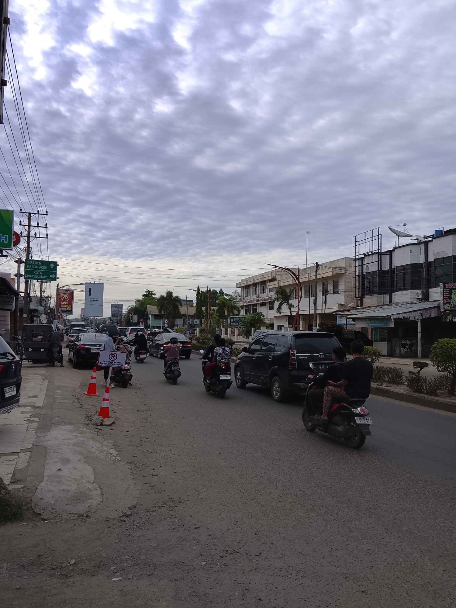 The image shows a street scene in Lhokseumawe, Indonesia. The road is busy with vehicles, including cars and motorcycles. There are several buildings lining the street, including shops and businesses. A road sign points towards "Bireuen Banda Aceh" and "Kota Lhokseumawe". A red and white sign reads "Dilarang Parkir Di Area Depan Apotek" indicating "No Parking in Front of Pharmacy". The sky is overcast with clouds. The road surface is asphalt, and there are some orange traffic cones on the side of the road. People are riding the motorbikes.