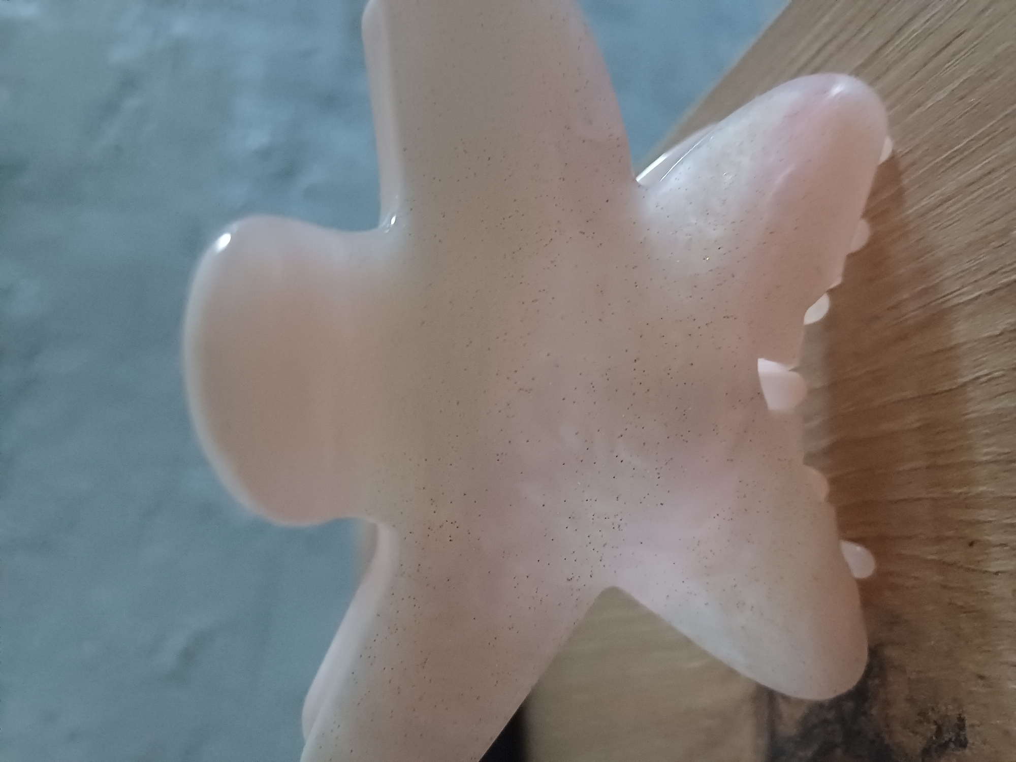The image shows a close-up of a pale pink, star-shaped hair claw. The claw has a slightly translucent appearance and contains small, dark flecks throughout its material. It is positioned on a light brown, wooden surface with a visible wood grain texture. In the background, a blurred, light blue wall is visible. The perspective is from a slightly elevated angle looking down at the hair claw. There are no people or animals present in the image, and no actions are depicted. The image was taken in Sulymivka, Ukraine.