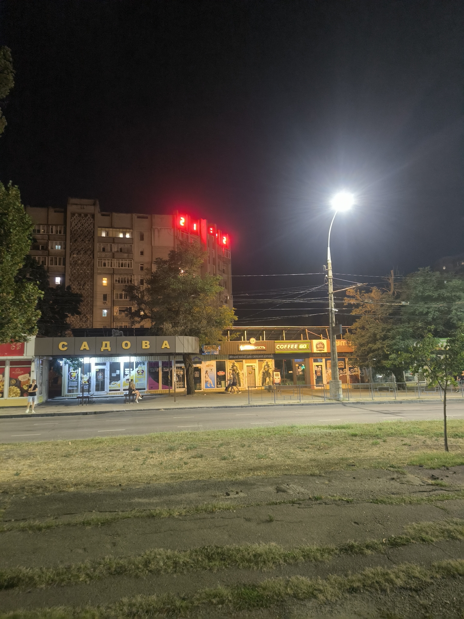 The image depicts a nighttime street scene in Mykolaiv, Ukraine, likely on Sadova Street, based on the prominent signage. A multi-story building with illuminated windows dominates the background. Red neon signs displaying the numbers "21" are visible on the upper facade of the building. In the foreground, several storefronts are illuminated. One storefront features yellow lettering spelling "САДОВА" which translates to "Sadova". Next to it, other businesses with signage including "COFFEE GO" and "VAPE SHOP" are visible. A street lamp with a bright bulb illuminates the scene from the right. A few people are visible walking or sitting on a bench outside the shops. The street in front of the buildings appears to be asphalt, with a grassy area on the left and a sidewalk. Trees are present on both sides of the buildings.