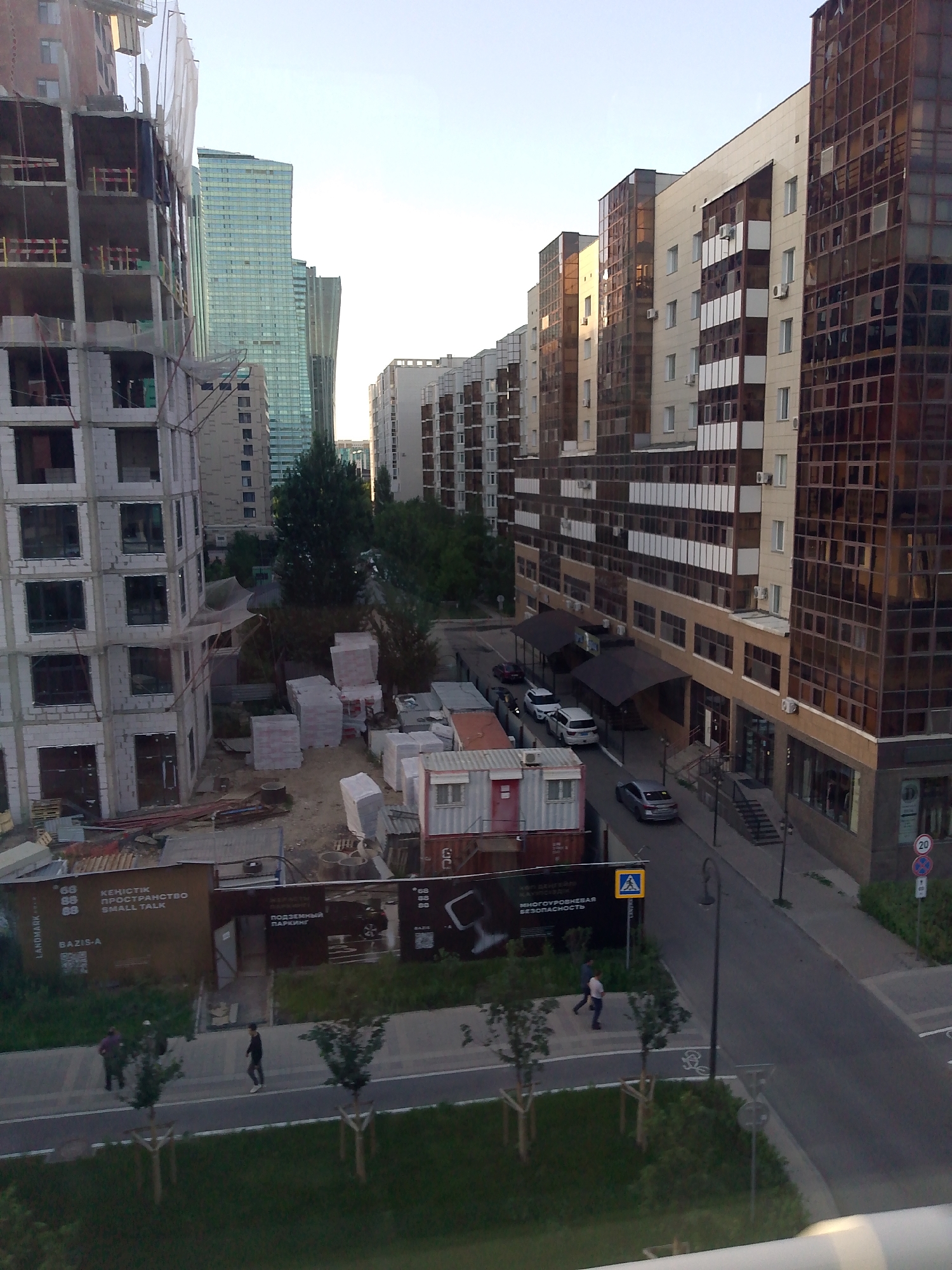This media file captures an urban scene in Astana, Kazakhstan, likely during the late afternoon or early evening due to the warm, diffused sunlight casting long shadows. The image features a mix of construction and completed modern buildings, suggesting ongoing development in the area.
In the foreground, a building under construction is prominent, with its concrete frame and exposed rebar visible. Construction materials, including stacks of bricks and modular units, are scattered around the site. Beyond the construction, a row of completed apartment buildings lines a street. These buildings are characterized by their contemporary architecture, with large windows and a combination of beige and dark-toned facades, some featuring reflective glass.
A street runs alongside the completed buildings, with several cars parked or driving. Pedestrians are seen walking on a sidewalk bordering the street, adding a sense of daily life to the scene. Overhead, the sky is clear and bright, indicating fair weather.
Visible text includes signs with Cyrillic script, such as "KEHICTIK ПРОСТРАНСТВО SMALL TALK" and "ПОДЗЕМНЫЙ ПАРКИНГ," along with brand names like "LANDMARK" and "BAZIS-A." Traffic signs, including a speed limit sign indicating "20," are also present. The overall impression is a dynamic urban environment undergoing growth and modernization.