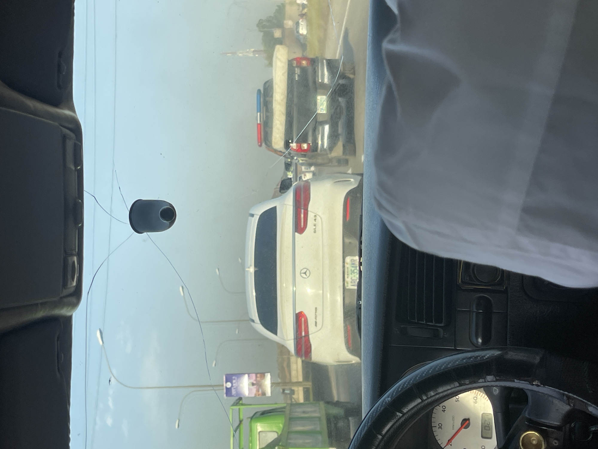 The image shows a view from inside a vehicle, looking out through a cracked windshield. In the foreground, parts of the dashboard and steering wheel are visible. In the midground, there is a white Mercedes-Benz GLE 43, followed by a black pickup truck carrying a mattress. Streetlights are also visible. In the background, the sky appears hazy. A billboard can be seen on the left, below the Mercedes. The license plate of the Mercedes reads "ARC-354MR" indicating that the image was taken in Jiru, Nigeria. The speedometer shows that the vehicle is moving at 20km/h.
