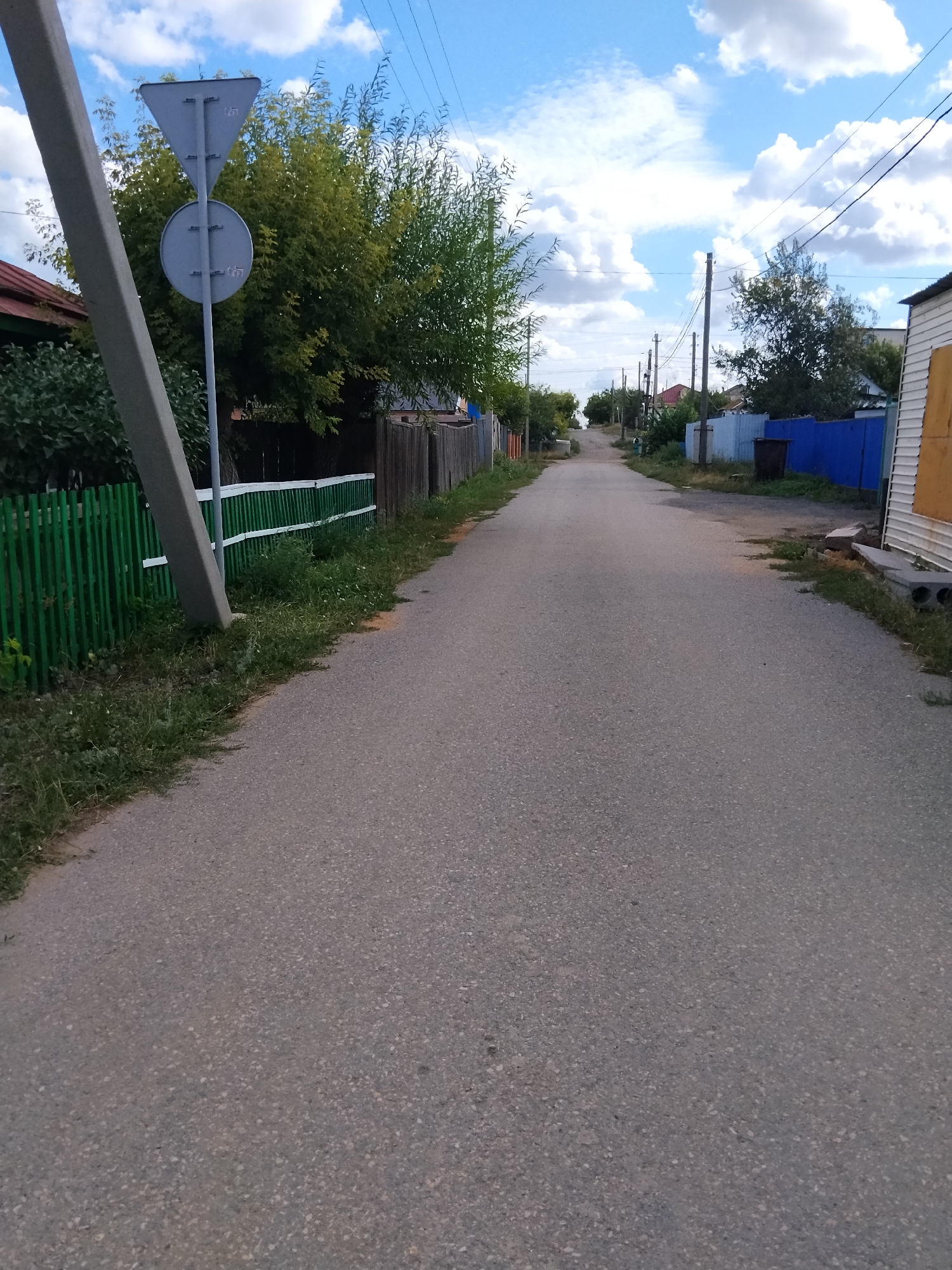 The image depicts a street scene in Kokshetau, Kazakhstan. The perspective is from ground level, looking down a paved road that appears to be in a residential area. On the left side of the road, there is a leaning concrete pole with a triangular sign and a circular sign attached. Further down the left side, a green wooden fence runs along the property line, bordered by lush green grass and some trees. On the right side of the road, there are houses with various fencing, including a blue fence and a wooden fence. A black trash bin is visible near the blue fence. Power lines are strung across the sky, supported by utility poles visible further down the street. The sky is blue with scattered white clouds. The overall impression is of a quiet, somewhat rural street on a clear day. No people are visible in the image.