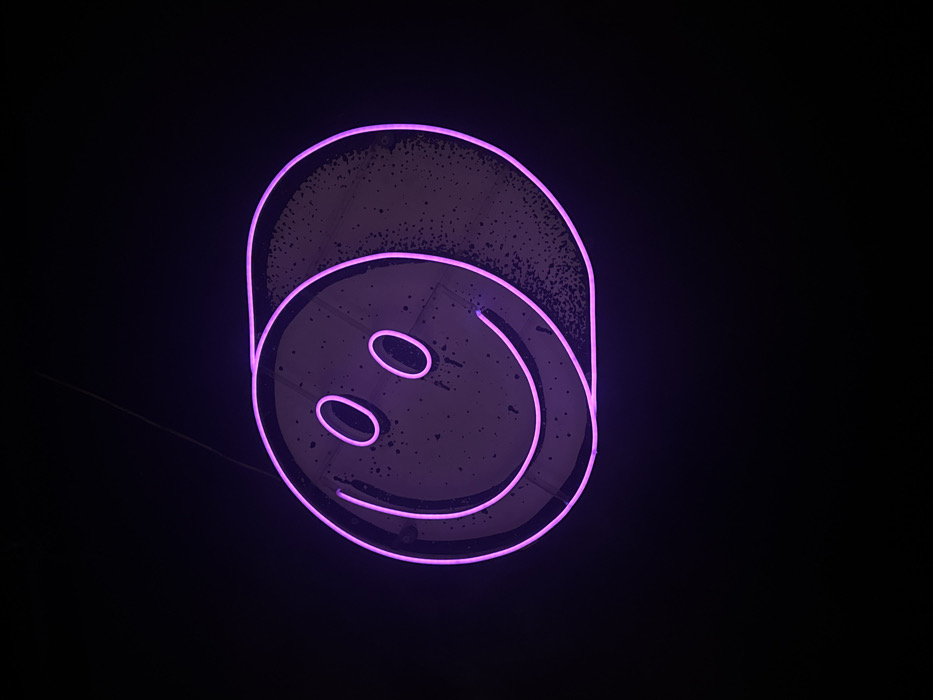 A magenta-purple neon sign, depicting a stylized smiley face, is observed against a dark, unidentifiable background. The sign is composed of glowing tubing forming a circular perimeter, two oval eyes, and an upward-curving mouth. Behind the illuminated neon elements, a circular panel, lighter in tone and marked with irregular dark spatters, is partially visible and illuminated by the neon light. A segment of this panel extends beyond the right side of the neon smiley's perimeter, forming a crescent-like shadow. A thin, dark suspension line is visible extending diagonally from the top edge of the frame toward the upper left portion of the sign. The scene is located in Tel-Aviv, Israel.