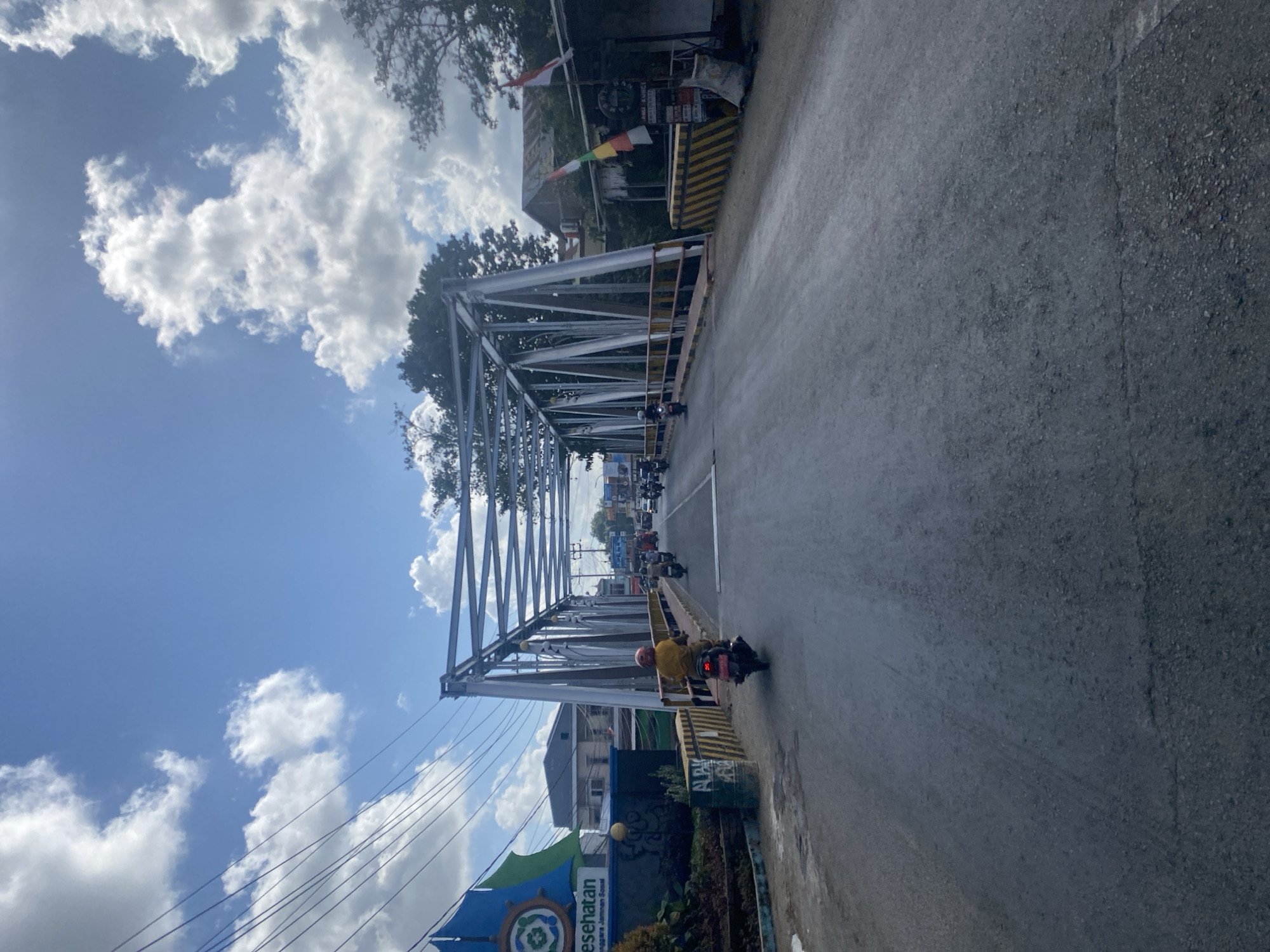 This image captures a view of a bridge in Bentengnge, Indonesia, under a partly cloudy blue sky. The bridge is a metal truss structure, with a road leading across it. A person on a motorcycle, wearing a yellow shirt and red helmet, is riding away from the camera towards the bridge. Several other motorcycles are visible on the bridge and in the distance, suggesting moderate traffic.
On the left side of the road, near the entrance to the bridge, there is a blue and green sign with text that appears to be in Indonesian, along with a circular emblem. The area surrounding the sign is landscaped with plants. Yellow and black striped barriers are present along the side of the road.
On the right side of the bridge, there is a building with flags, including the Indonesian flag, displayed. The environment appears to be urban or semi-urban, with buildings and trees visible in the background. The road surface is asphalt and shows some wear. Power lines are strung overhead, running parallel to the road and bridge.