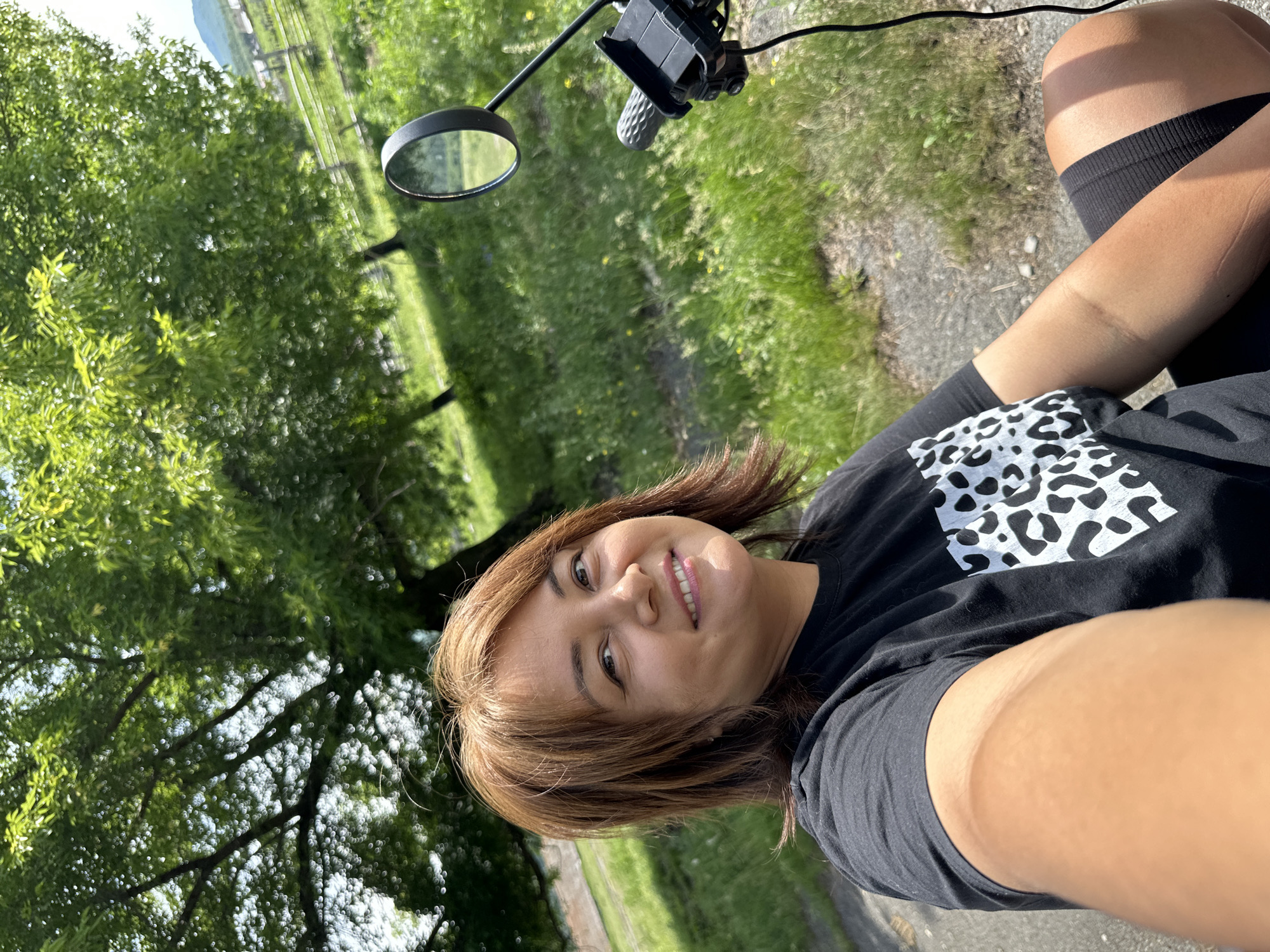 A selfie is taken outdoors on a sunny day. The subject, a woman with brown hair and a black t-shirt with a white leopard print pattern, is looking at the camera and smiling. She is sitting on what appears to be a motorcycle, with a black handlebar and a rearview mirror visible to her right. The background consists of lush green grass, trees, and a wooden fence. The overall scene suggests a rural or park-like setting.