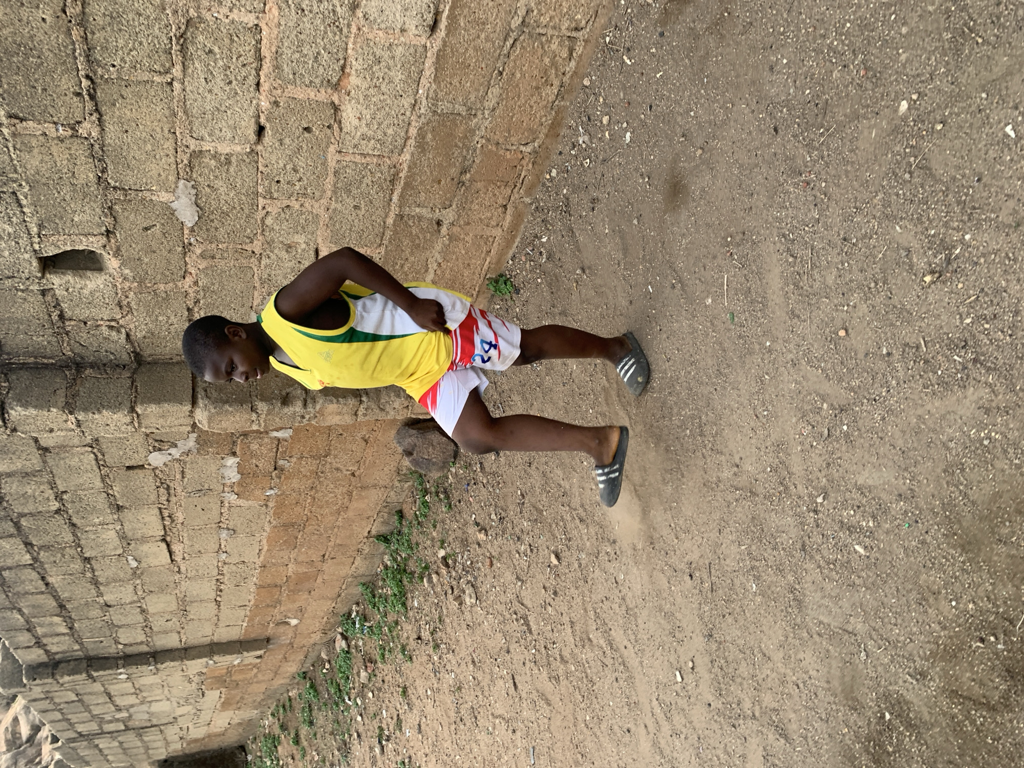 A young male is standing on a dirt surface next to a brick wall. He is wearing a yellow and white tank top and red and white shorts. He is also wearing black sandals with white stripes. The dirt surface is light brown in color and appears to be a mix of sand and small stones. There are some green plants growing at the base of the brick wall. The location is Jos, Nigeria, based on visual context.