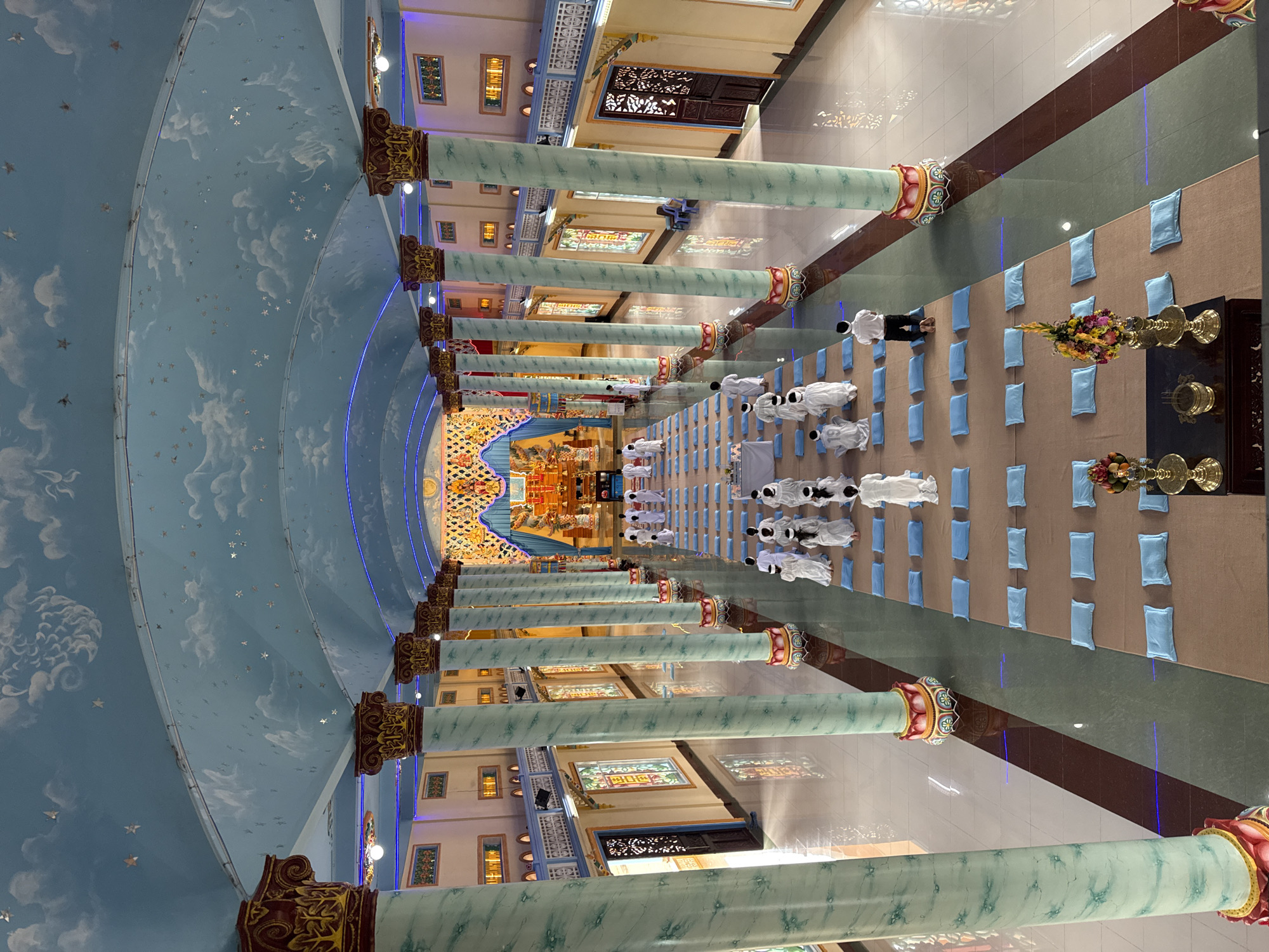 The image captures the interior of a religious building, possibly a temple or monastery, in Lộc Quý, Vietnam. The building's architecture includes a sky-blue, cloud-painted arched ceiling with star accents, supported by rows of pale green columns with ornamented capitals. Along the floor, individuals in white robes are kneeling on blue cushions in orderly rows. At the far end, an ornate altar with various religious objects is visible. The floor is covered with sections of brown and green carpeting. The scene suggests a ritual or religious service taking place.