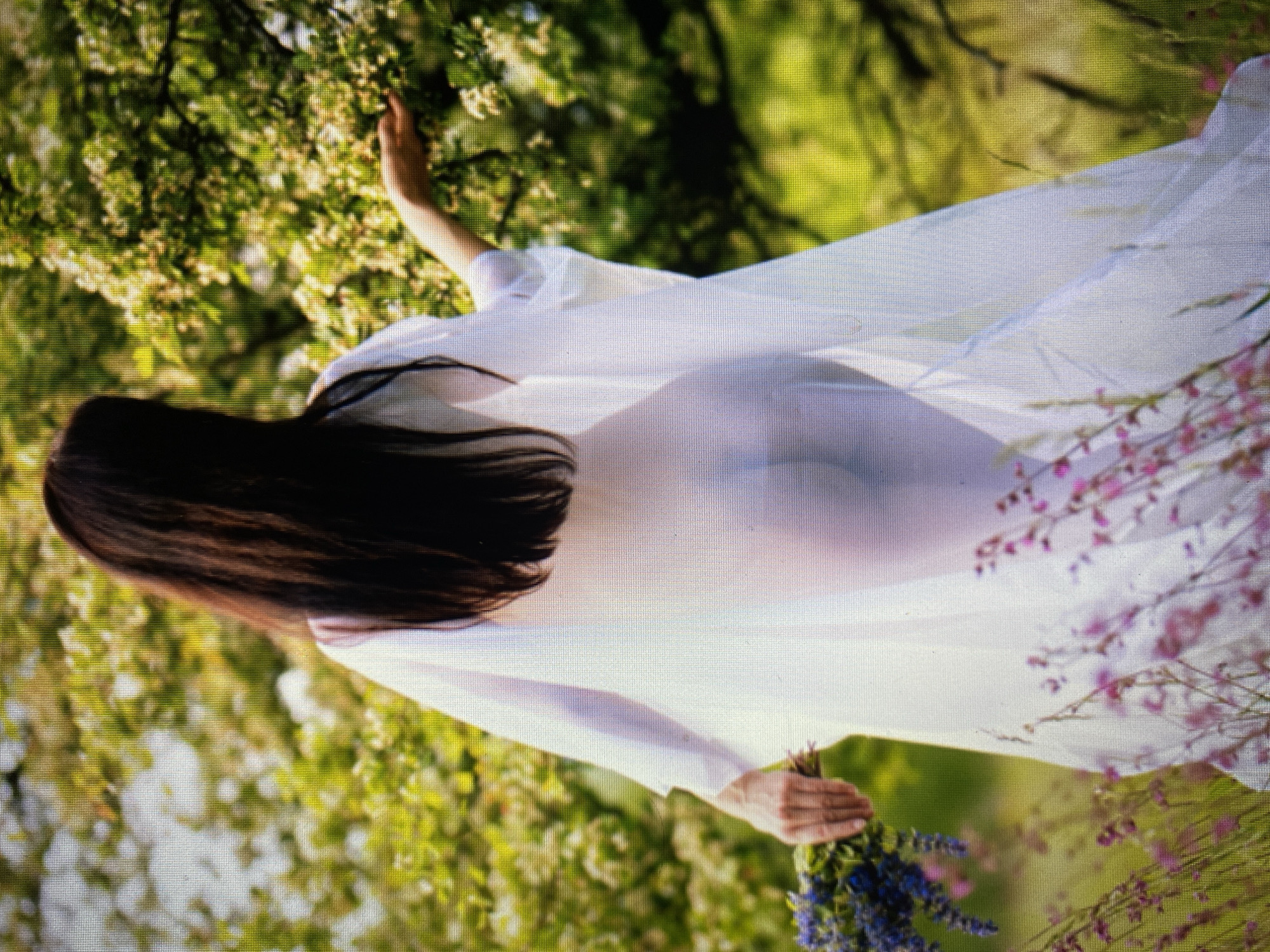 A woman with long dark hair, wearing a sheer white dress, stands in a field of green grass and flowers. She is holding a bouquet of purple flowers in her left hand, and her right hand is extended towards a branch of a flowering tree. The dress is translucent, revealing the woman's body underneath. The background is a blur of green foliage and white blossoms, suggesting an outdoor setting in a natural environment. The lighting is soft and diffused, creating a dreamy atmosphere.