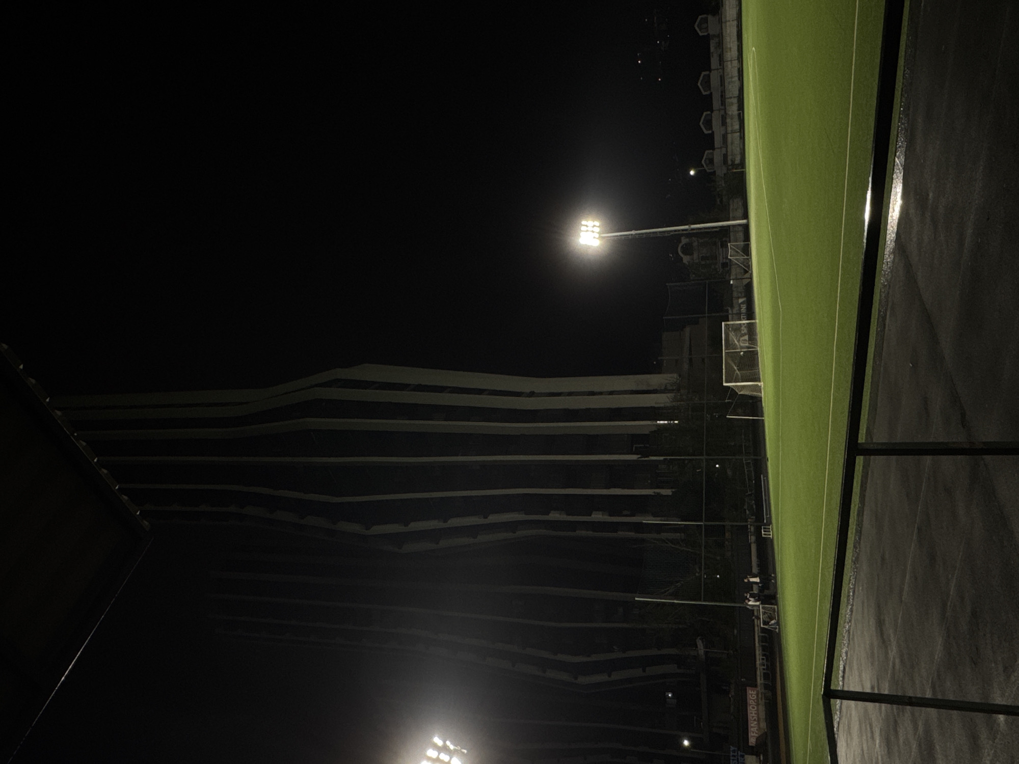 The image depicts an illuminated artificial turf field at night in Tbilisi, Georgia. The view is oriented so that the field runs along the right edge of the frame. The field is surrounded by dark surfaces. A tall building is located across the street. The sky is obscured by darkness. A lighting tower is visible above the field. Two people are standing on the field by a goal.