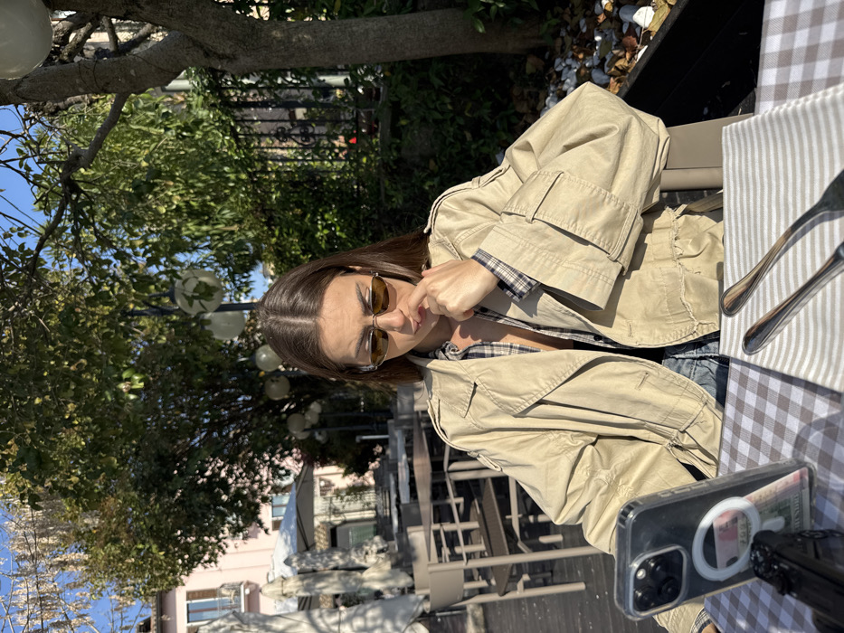 A lone female individual is depicted seated outdoors at a cafe table within the historic center of Sirmione, Italy. The background features a light pink building with balconies to the left, and multiple green-leafed trees adorned with spherical decorative lights. The woman has medium-long brown hair and wears small, brown-tinted oval sunglasses, a beige trench coat over a partially unbuttoned plaid shirt, and blue denim. Her right index finger rests against her chin as she gazes downward. On the brown and white checkered tablecloth, silverware (a fork and knife) is positioned on a striped napkin. A smartphone in a clear case, with a MagSafe-style ring and displaying a currency note, is propped upright. A small black object rests beside the phone. The scene is brightly illuminated by direct sunlight, indicating a clear day.