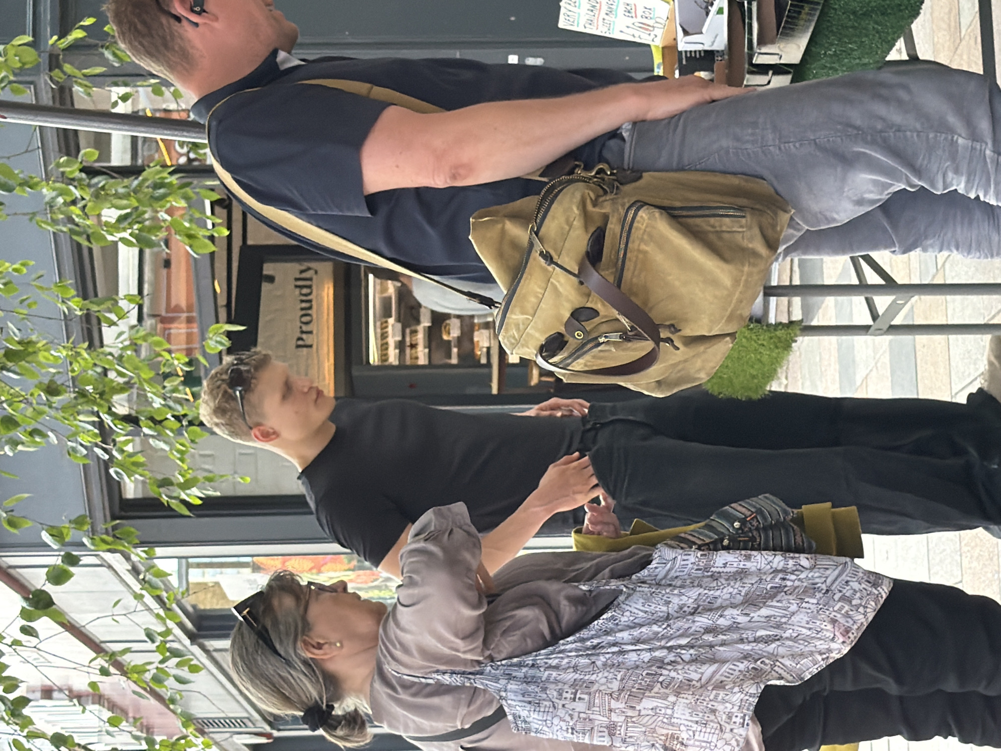 Three people are shown in this outdoor scene, which appears to be a street market. The woman in the foreground on the left, with grey hair pulled back in a ponytail and wearing glasses, has a printed tote bag slung over her shoulder. To her right, a young man with blond hair and sunglasses on his head stands facing away from the camera. He is wearing a black t-shirt and dark trousers. The third person, a man on the right, faces left and is wearing a navy blue shirt and grey trousers. A canvas messenger bag with brown leather accents is slung across his body. Behind the people, a storefront with a sign reading "Proudly" is visible, suggesting a commercial area, possibly within a city. The ground is paved with cobblestones, and greenery hangs from above, hinting at an urban setting. The overall atmosphere is casual and suggests a daytime activity.