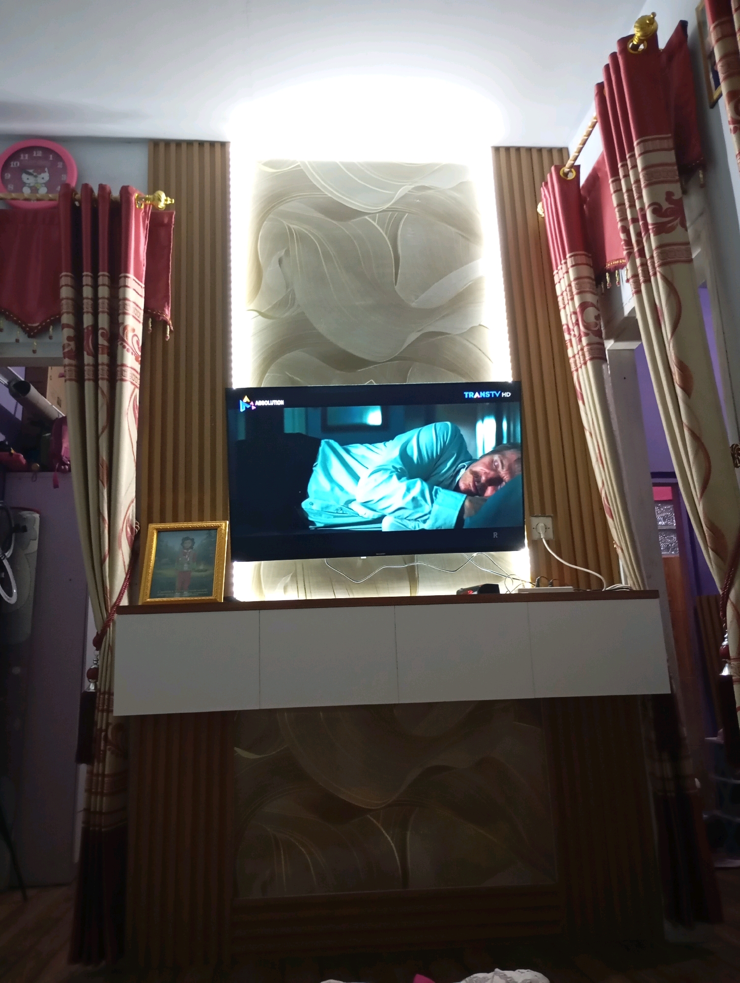 The image shows a living room scene in Muncang, Indonesia, featuring a television displaying a man lying in bed, and red and gold curtains on either side of the screen. A golden framed picture sits on a white cabinet below the TV. The room is decorated with vertical wooden panels on the walls. The overall ambiance appears to be domestic and relaxed.