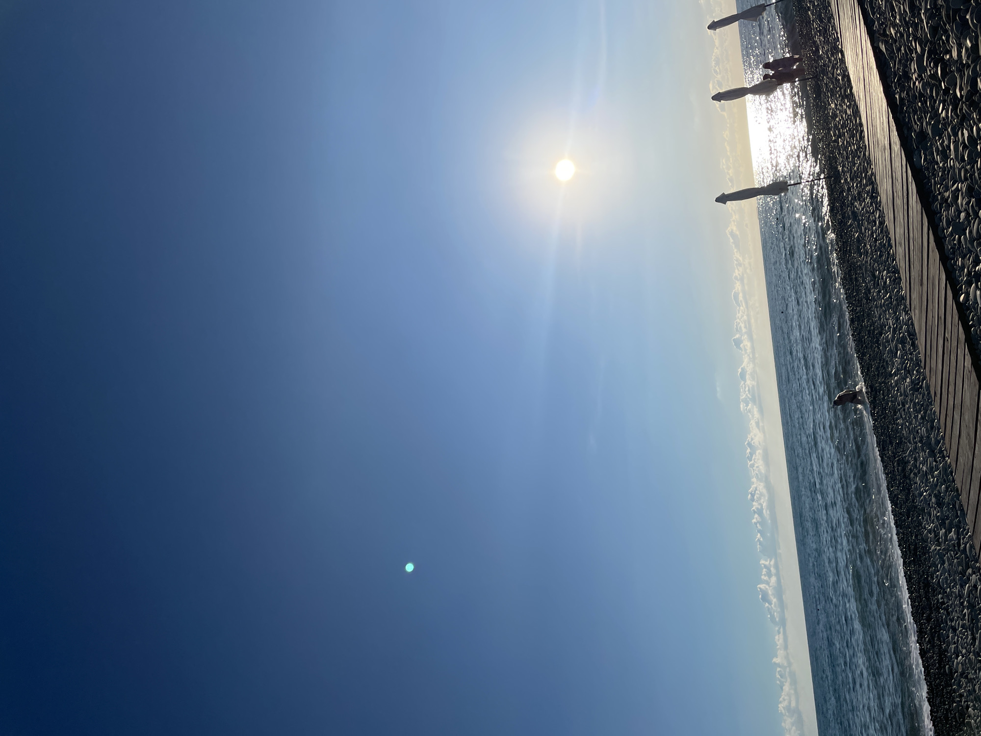 The image depicts a serene beach scene with a clear blue sky dominating the upper portion. The sun is brightly shining, positioned in the mid-right of the frame, casting a luminous reflection on the ocean's surface. Gentle waves are lapping against a pebble beach, which is partially covered by a wooden walkway in the foreground. Several beach umbrellas, closed and standing upright, are scattered along the shoreline. In the distance, a solitary person is wading in the shallow water, while a small group of people are visible near the umbrellas. The horizon is marked by a line of soft, white clouds. The overall atmosphere is peaceful and suggests a pleasant day at the beach.