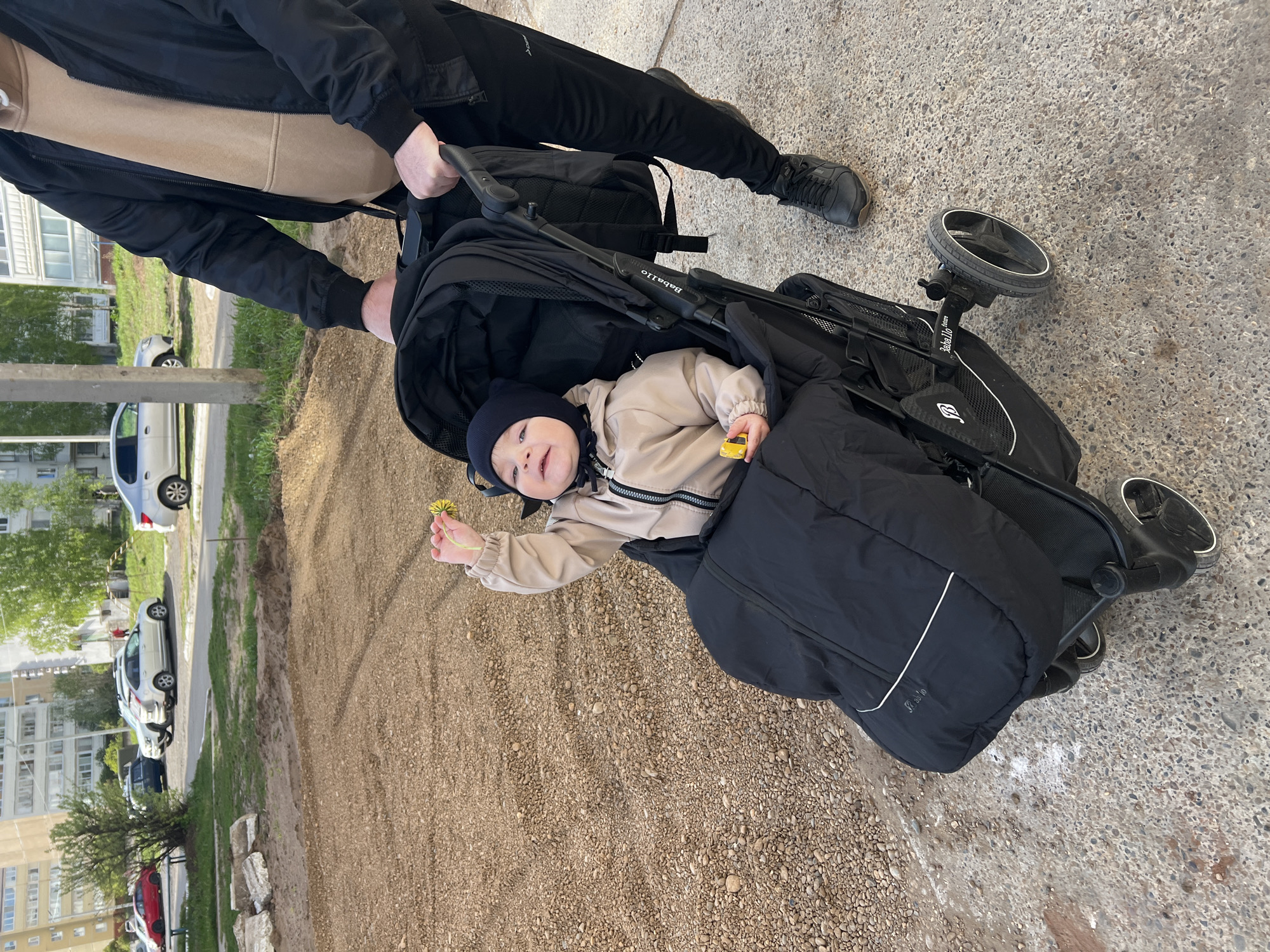 A baby wearing a navy blue hat and a light-beige jacket, lying in a black Boballo stroller, is holding a small yellow flower. The baby's stroller is being pushed by an individual dressed in black. The location appears to be Noviy, Russia, with a gravel surface visible adjacent to a paved path. Cars and apartment buildings are visible in the background. The surface of the ground around the stroller is divided between a paved path and the gravel-covered section. The image is oriented sideways.