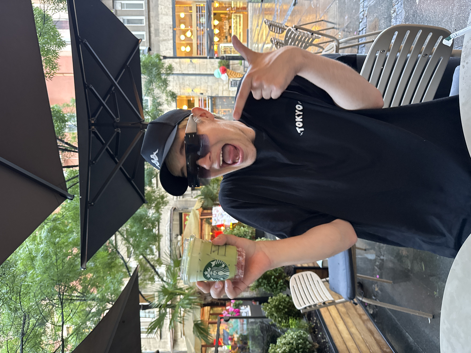 A person wearing a black "TOKYO" t-shirt, black baseball cap, and sunglasses is holding a Starbucks cup with a green drink. The person is gesturing with their right hand and has their mouth open. The scene is outdoors, possibly a cafe in the city of Belgrade, Serbia. A black umbrella is partially visible. The background features buildings, greenery, and outdoor seating.